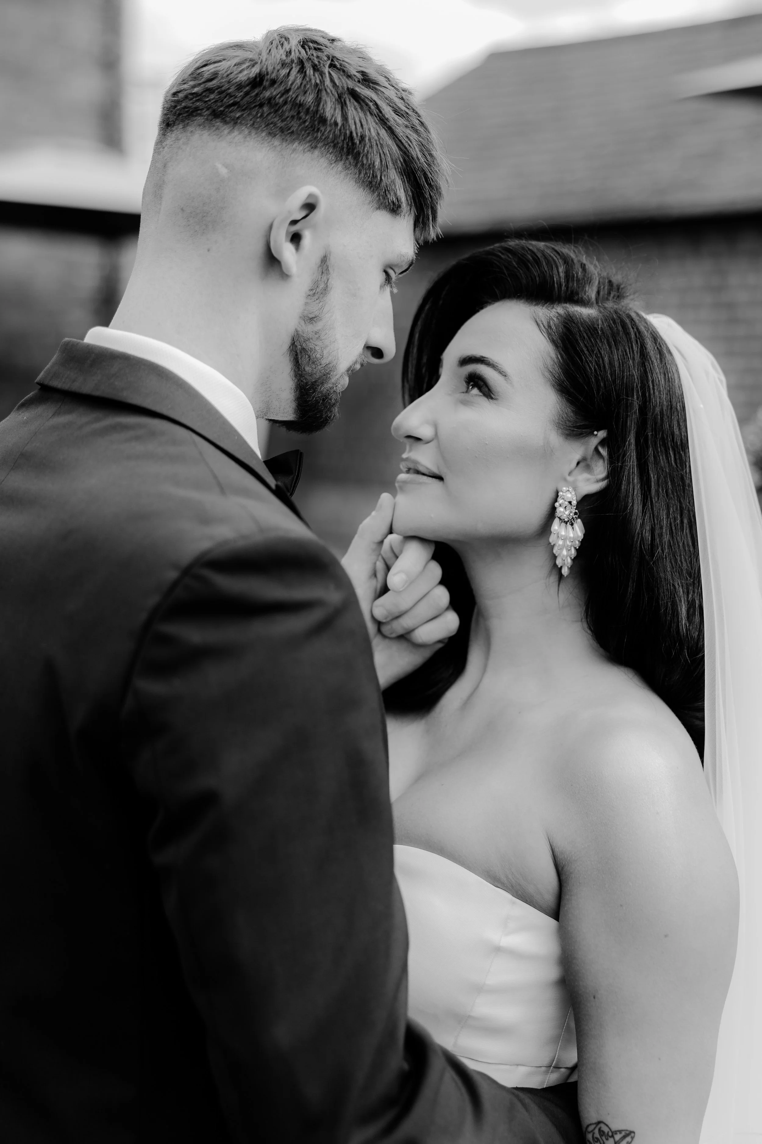 A black-and-white photo of a bride and groom looking into each other's eyes, with the groom gently touching the bride's chin, during their wedding.