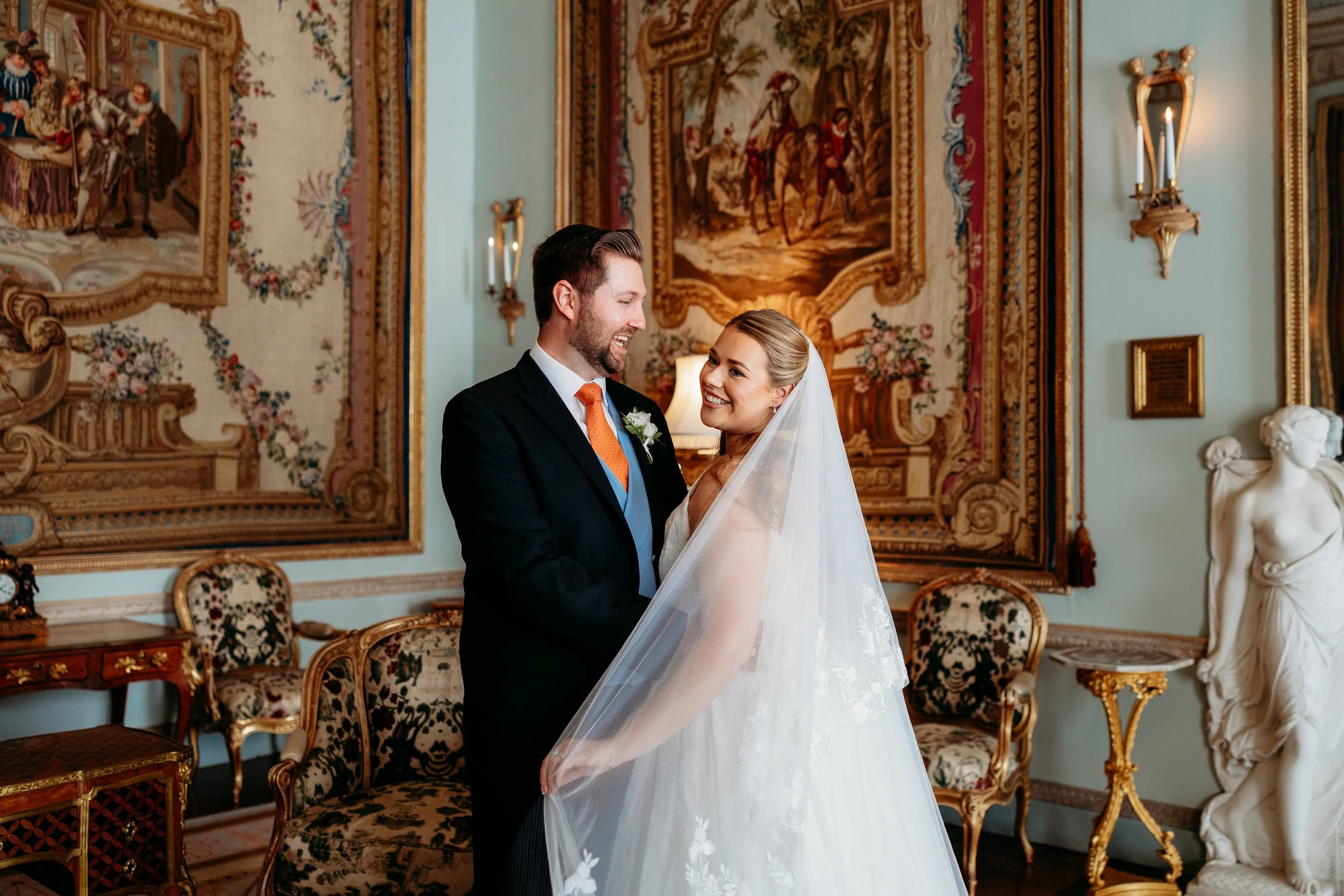 A newlywed couple sharing a joyful moment during their wedding in an elegant, ornate room with antique furnishings and large, elaborate tapestries at Goodwood Estate, Sussex.