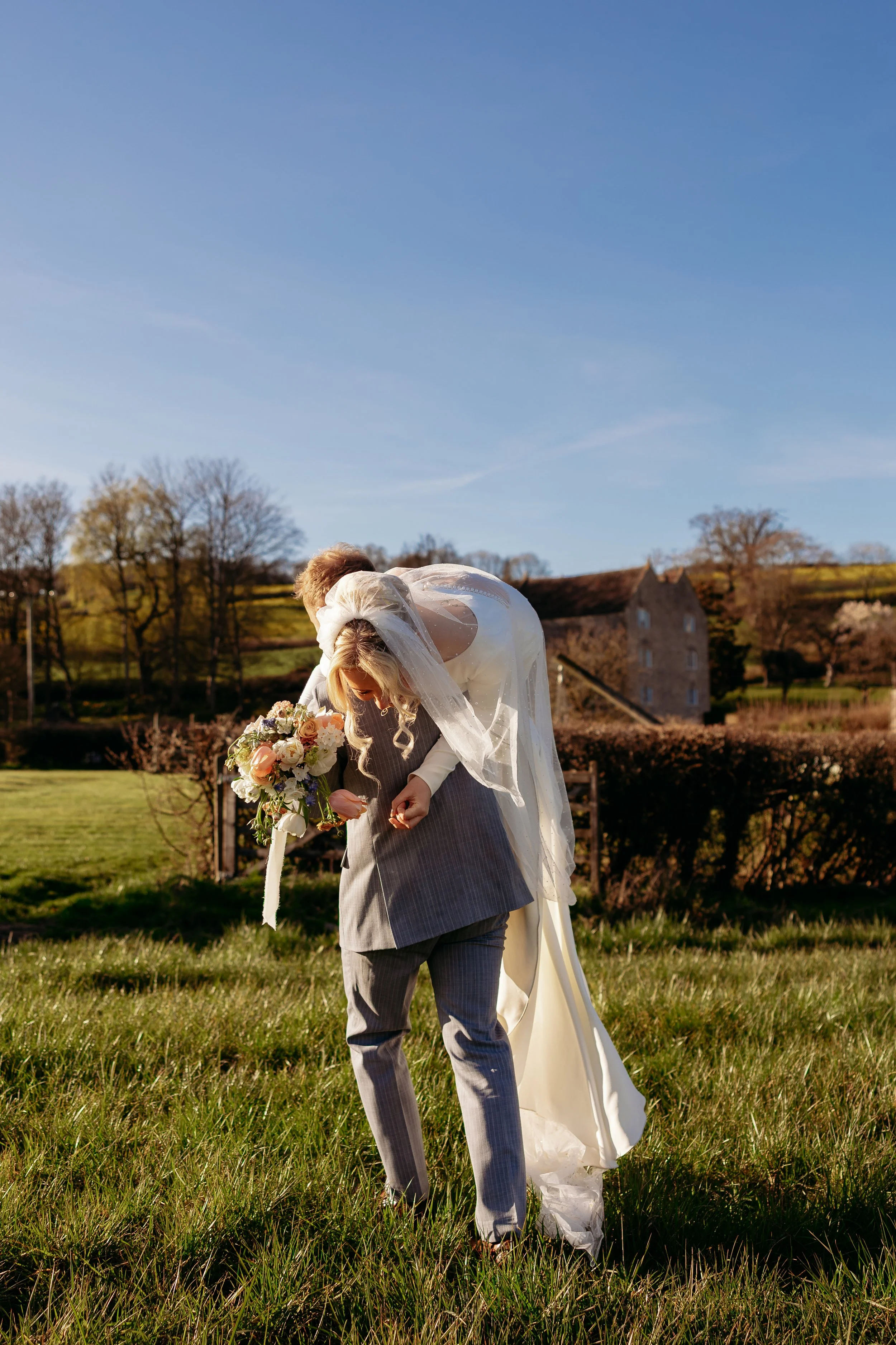 Bride and groom standing outdoors on grass, with the bride holding a bouquet, during a ski. The bride leans forward, resting on the groom's back, under a clear blue sky with trees and a building in the background.