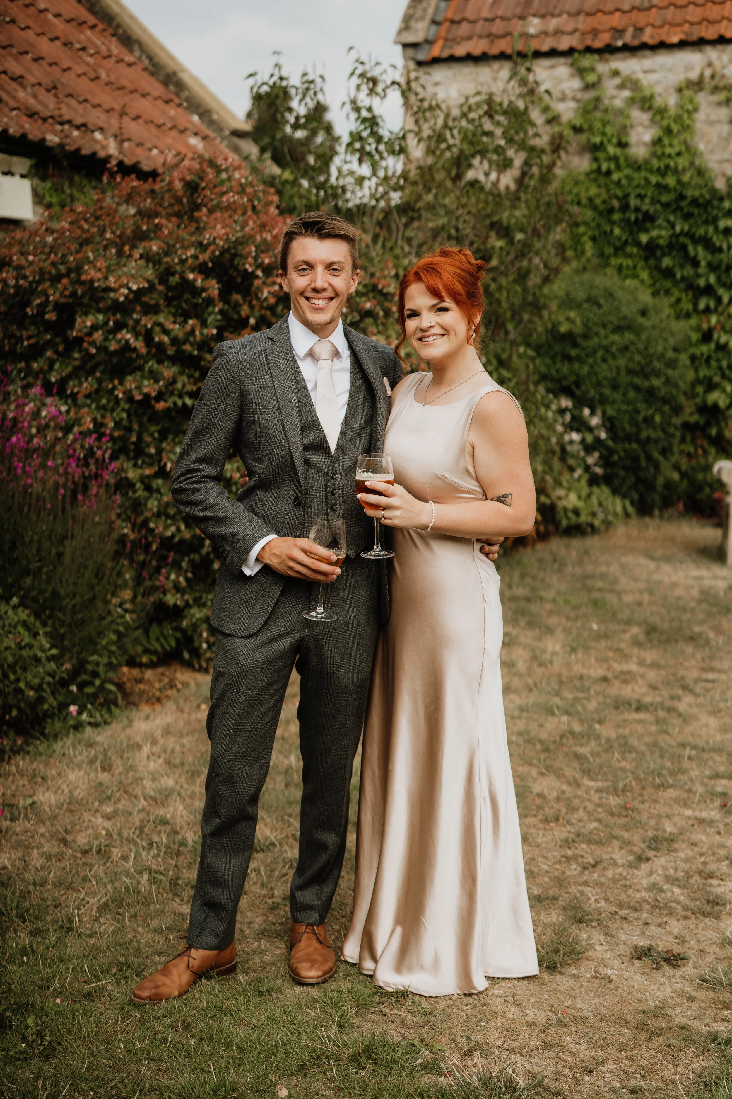 A man and woman dressed in formal attire smiling and holding drinks outdoors near bushes and trees, with buildings and cloudy sky in the background.