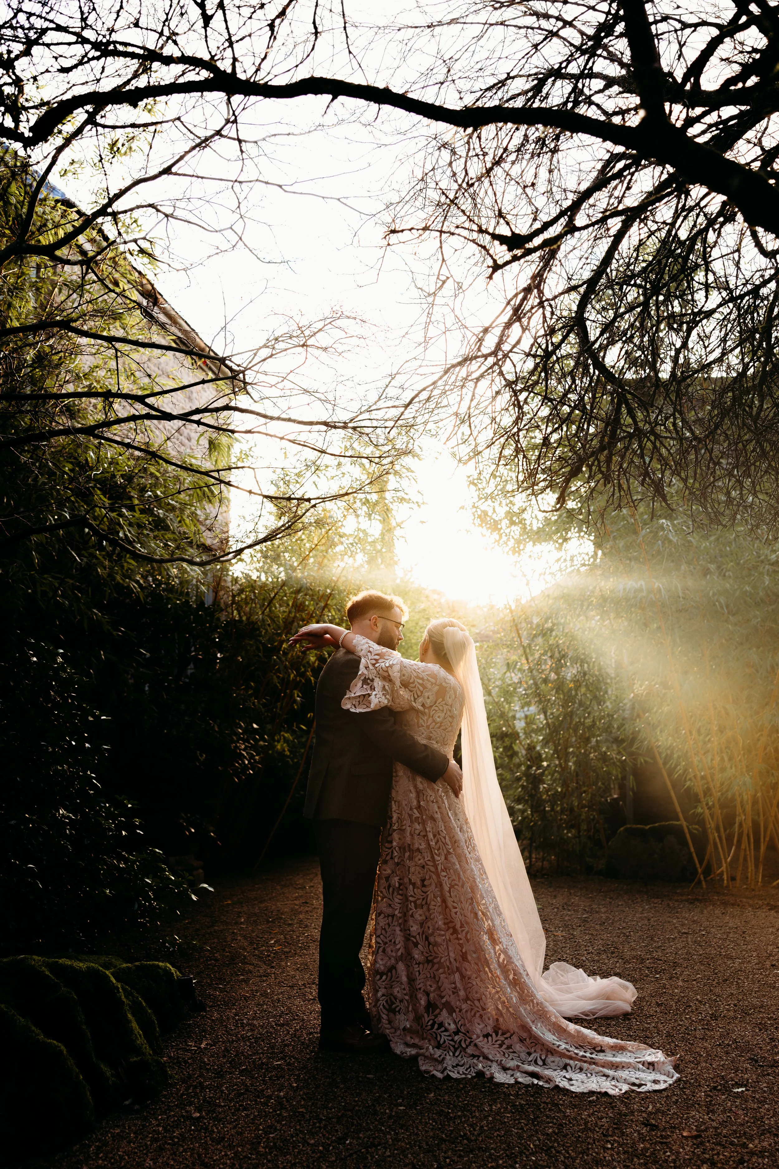 A bride and groom embracing outdoors during sunset, surrounded by trees and greenery, with sunlight filtering through branches.