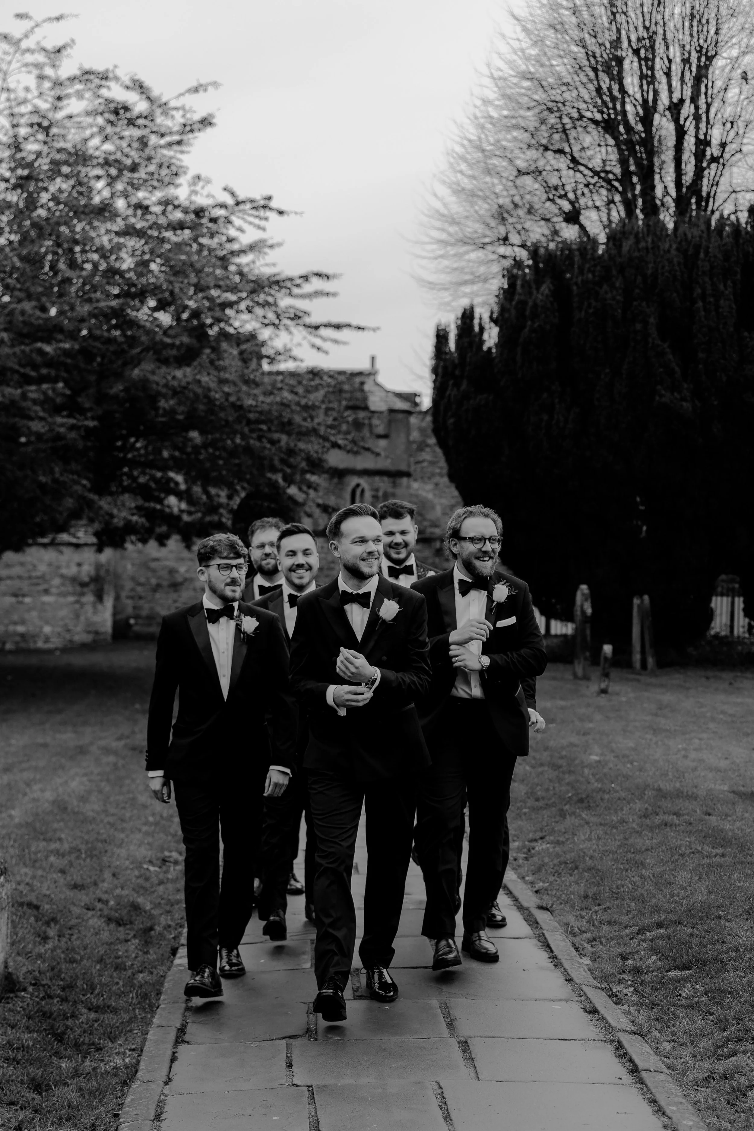 A group of six men in tuxedos walking together outdoors on a stone path, smiling and enjoying a formal event, with trees and buildings in the background, in black and white.