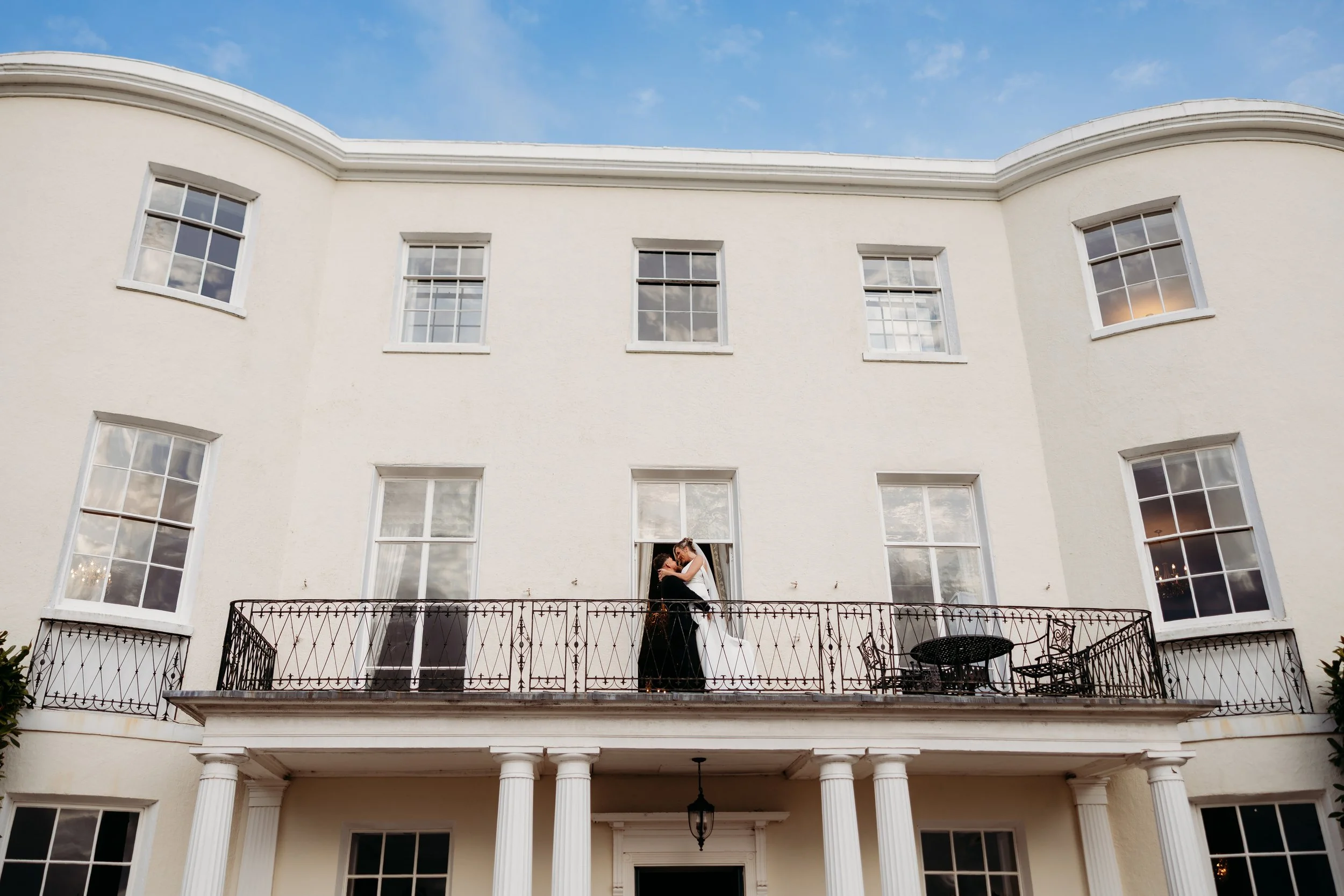 Couple embracing on a balcony in front of a large white building with multiple windows and columns, under a blue sky with some clouds.