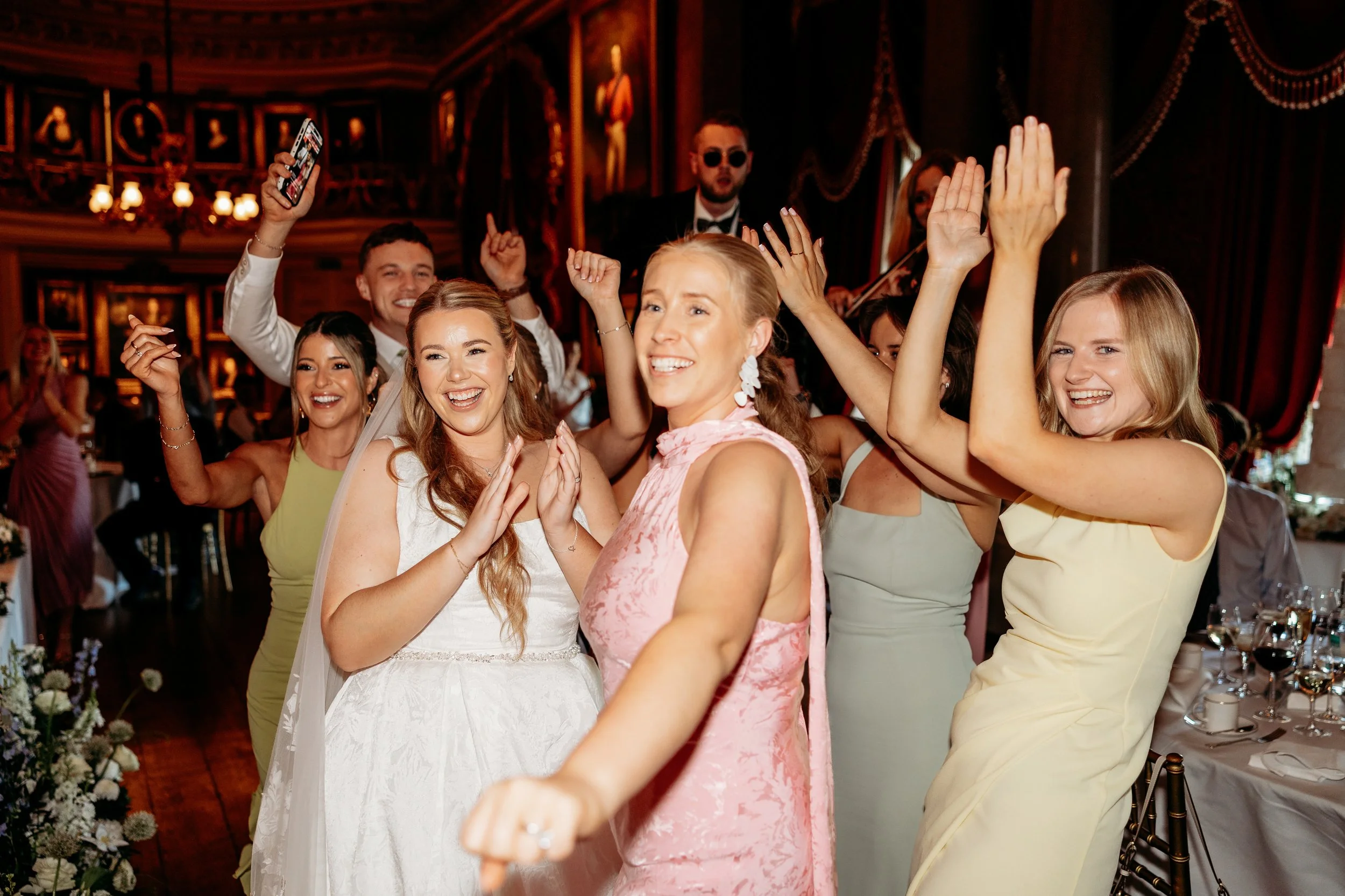 Group of people celebrating at a wedding reception, smiling, clapping, and raising their hands, with a bride in white dress and others in formal attire at Goodwood Estate, Sussex.