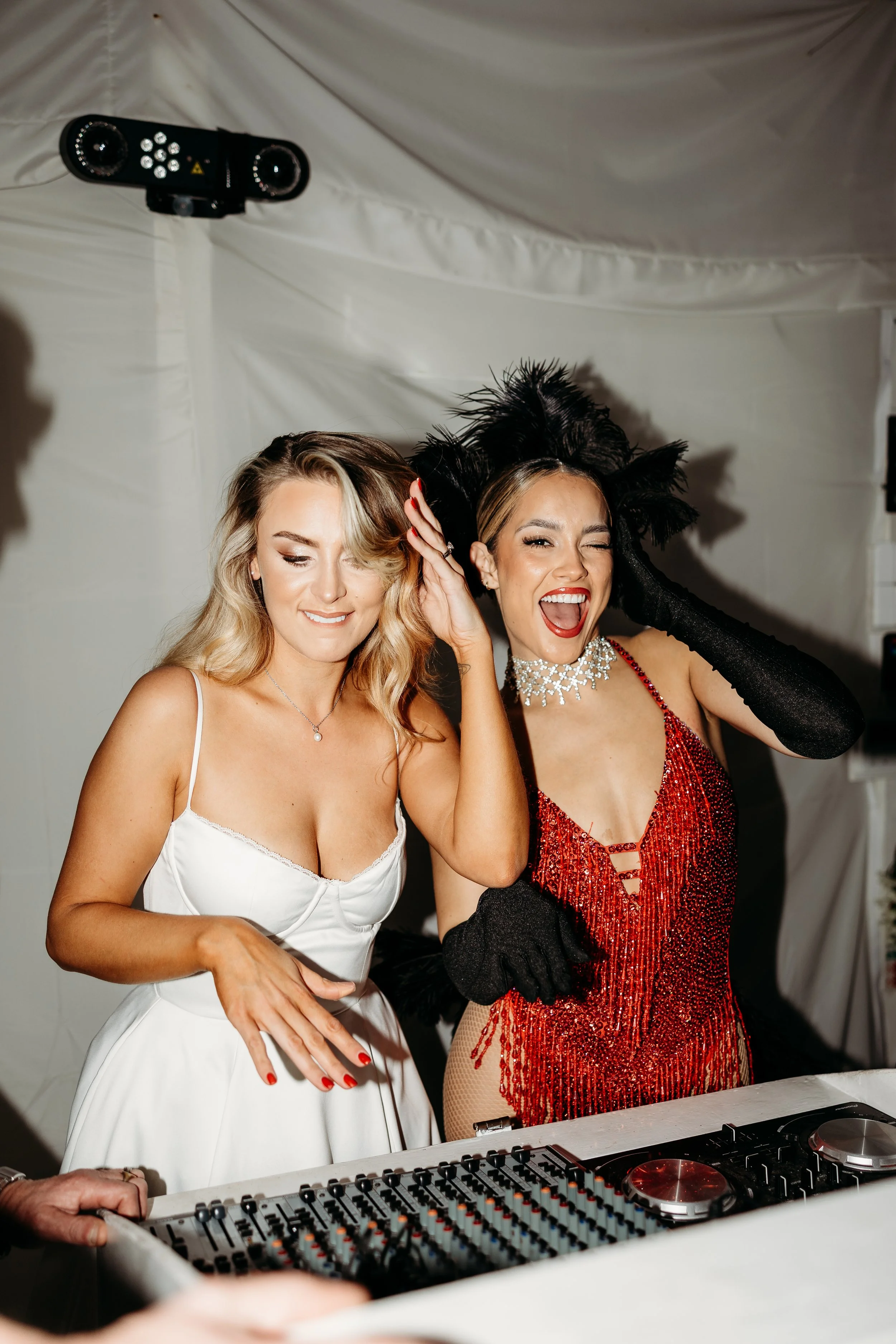 Two women dressed glamorous, one in a white dress and the other in a red sequin dress, enjoying music at a party with DJ equipment in front of them. A fun wedding evening.