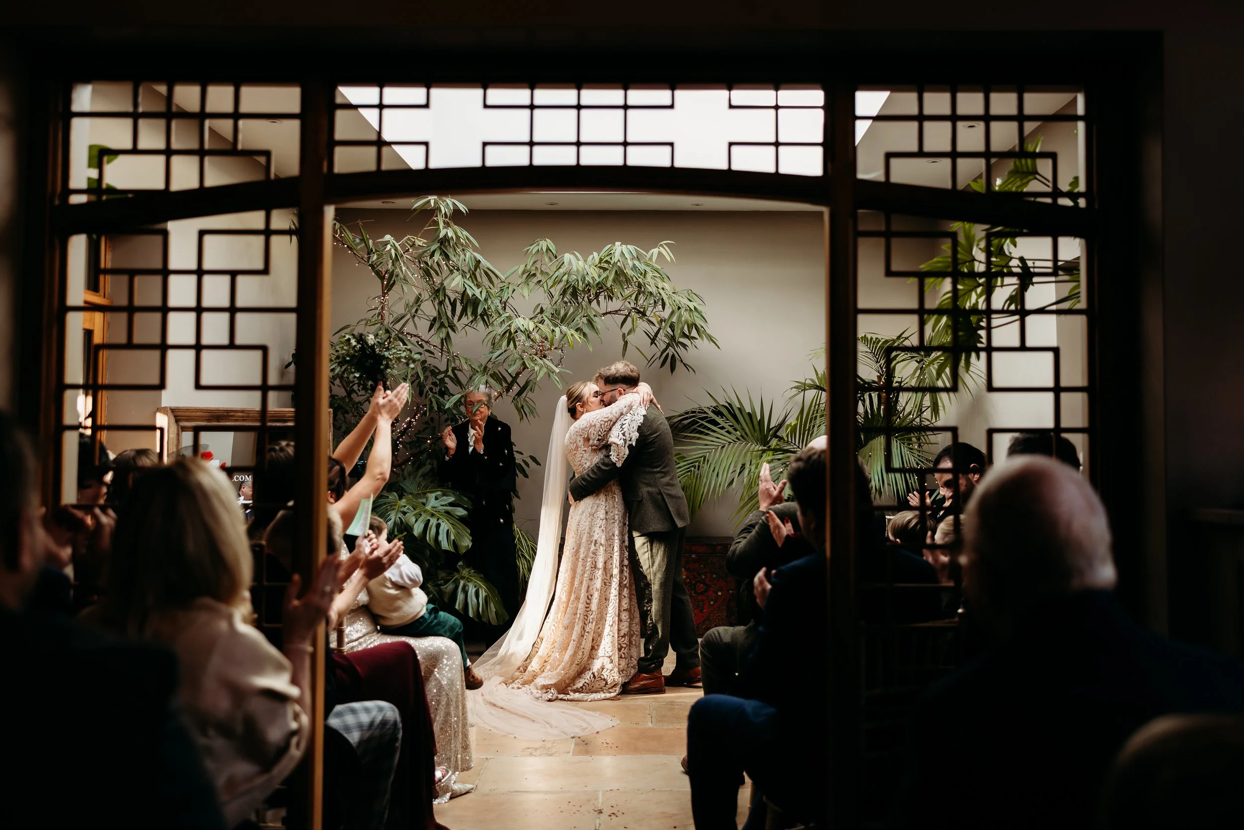 A wedding ceremony with a bride and groom embracing and kissing, surrounded by guests clapping, in a room with large tropical plants at Matara Centre, Cotswolds.
