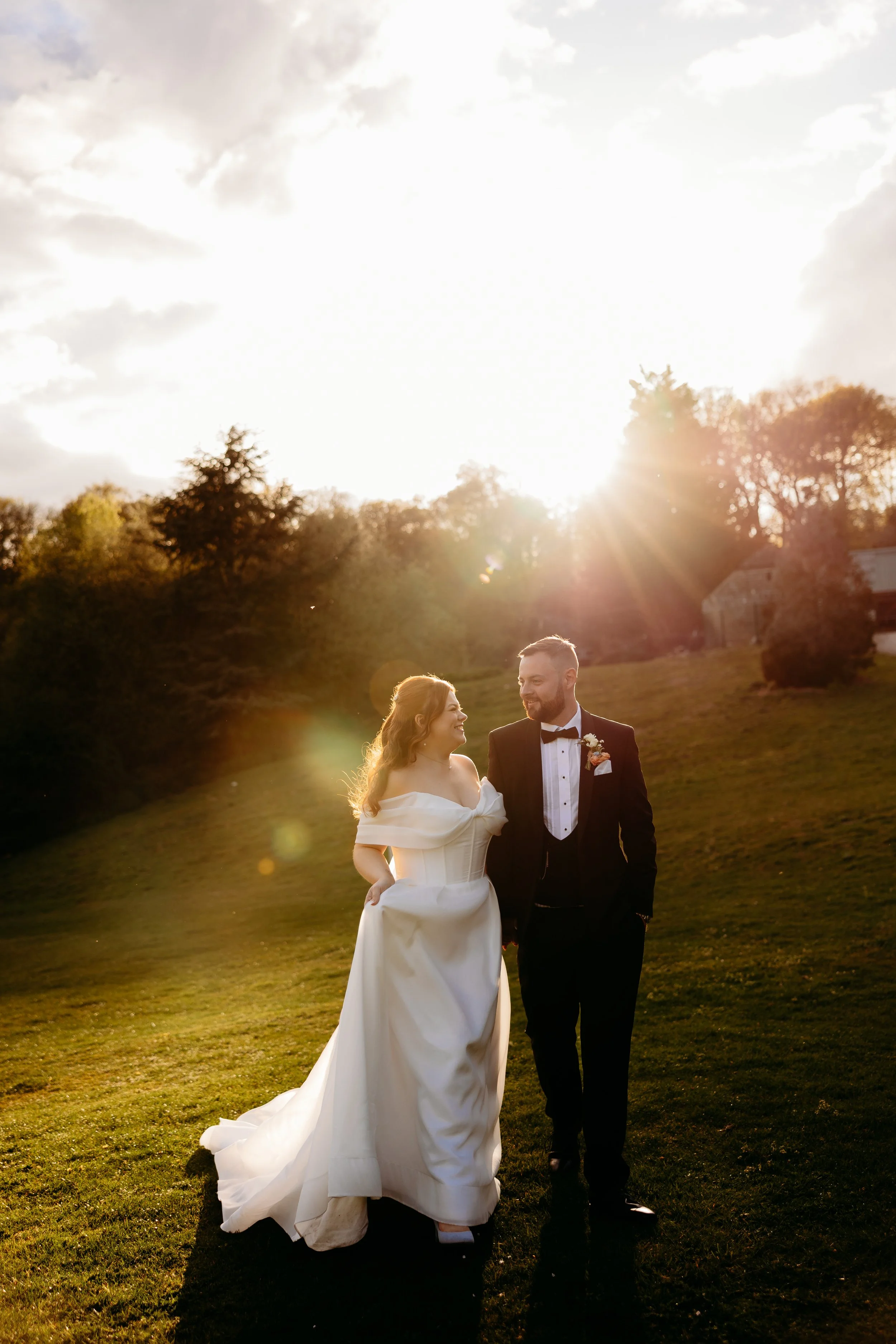 A bride and groom walking outdoors in sunlight, with trees and a house in the background.