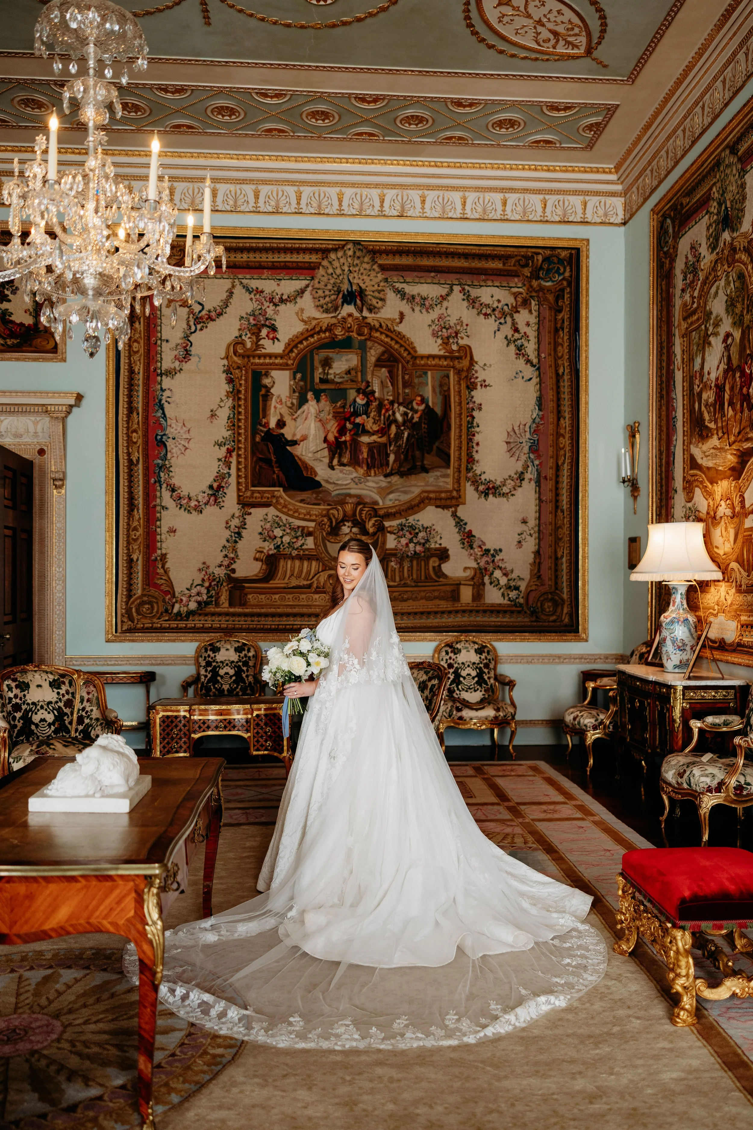 A bride in a white wedding gown with lace details, holding a bouquet of white flowers, standing in an ornate room with antique furniture, elaborate tapestries, a large chandelier, and intricate ceiling decorations at Goodwood Estate, Sussex.