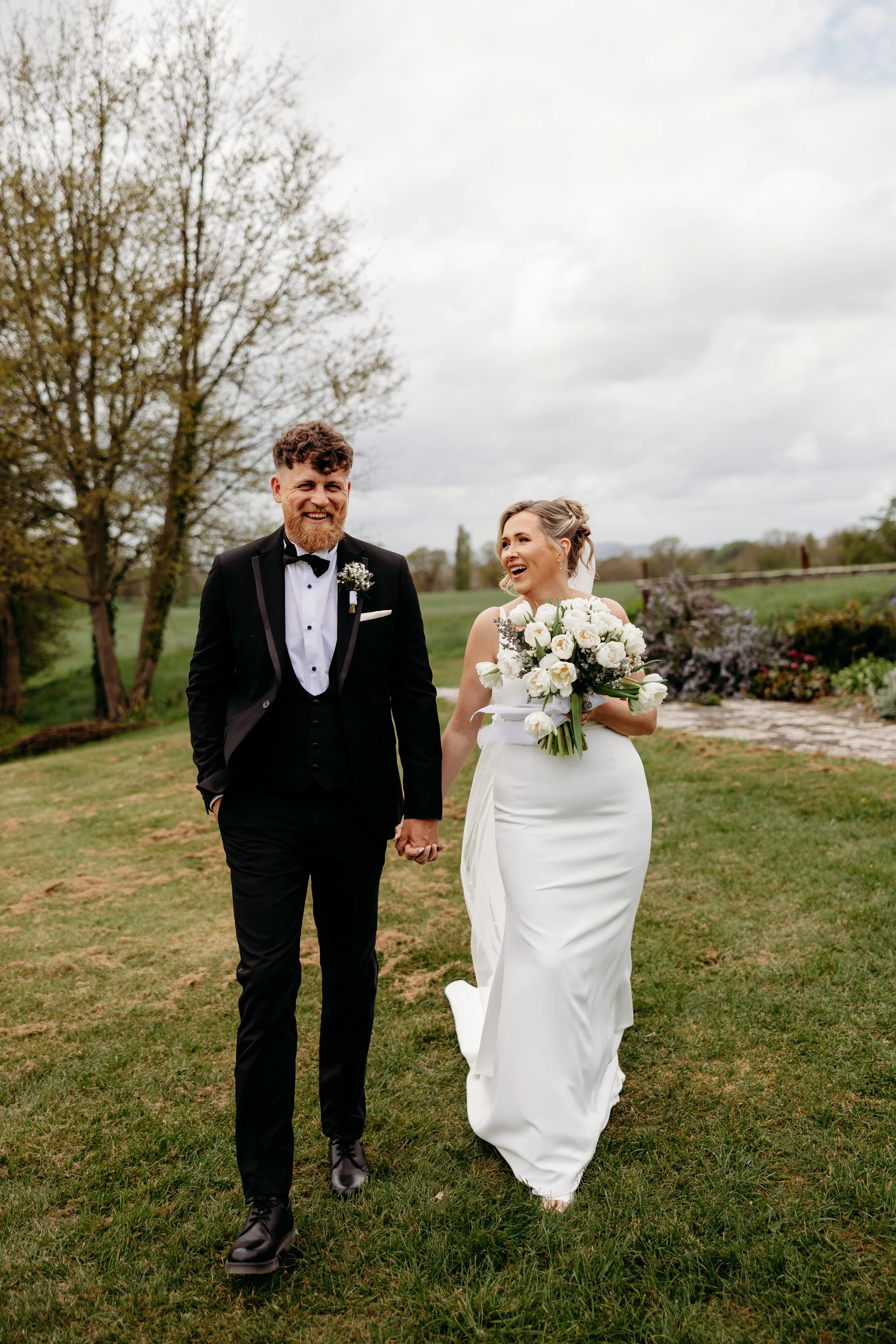 A couple in wedding attire walking outdoors on a grassy area during daytime, holding hands and smiling, with trees and a cloudy sky in the background.