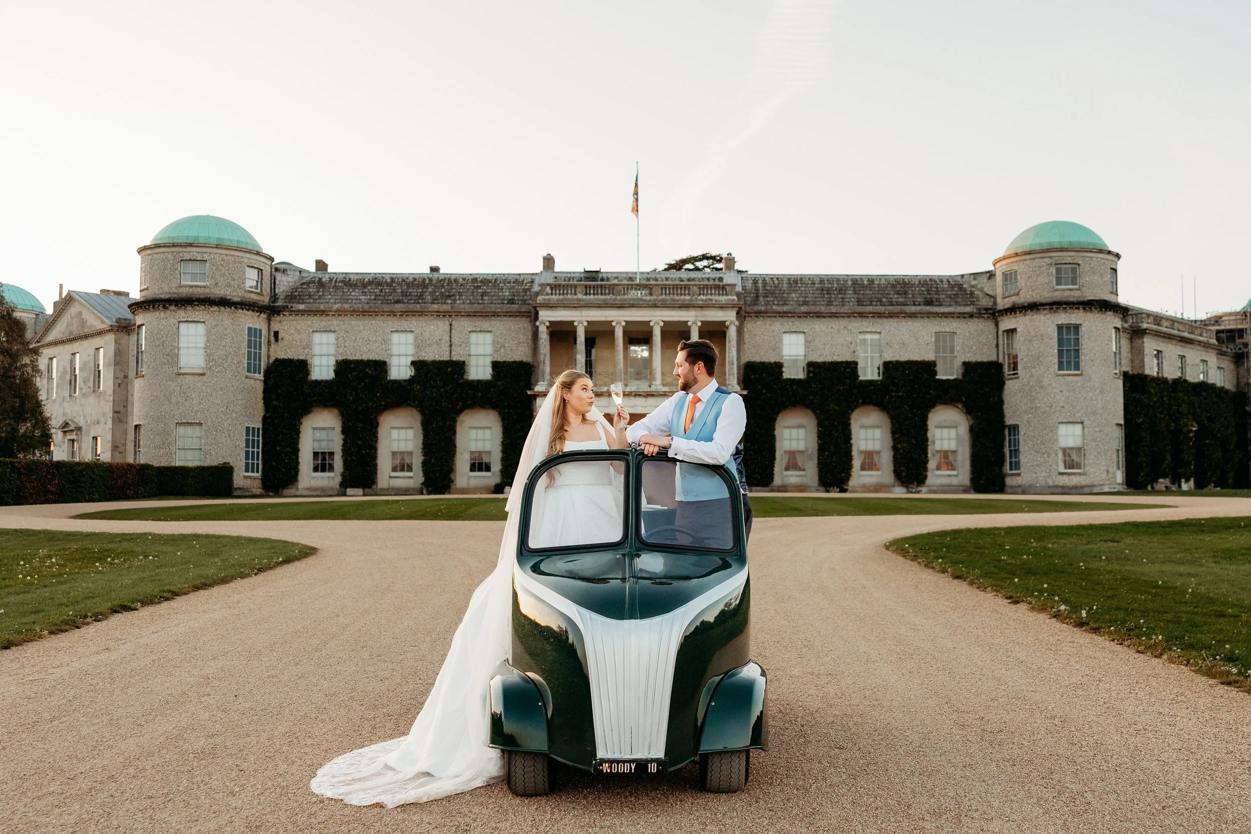 A bride in a wedding gown and a groom in formal attire stand inside a small vintage car with the bride holding a champagne glass, in front of a historic mansion with green domed roofs at Goodwood Estate, Sussex.
