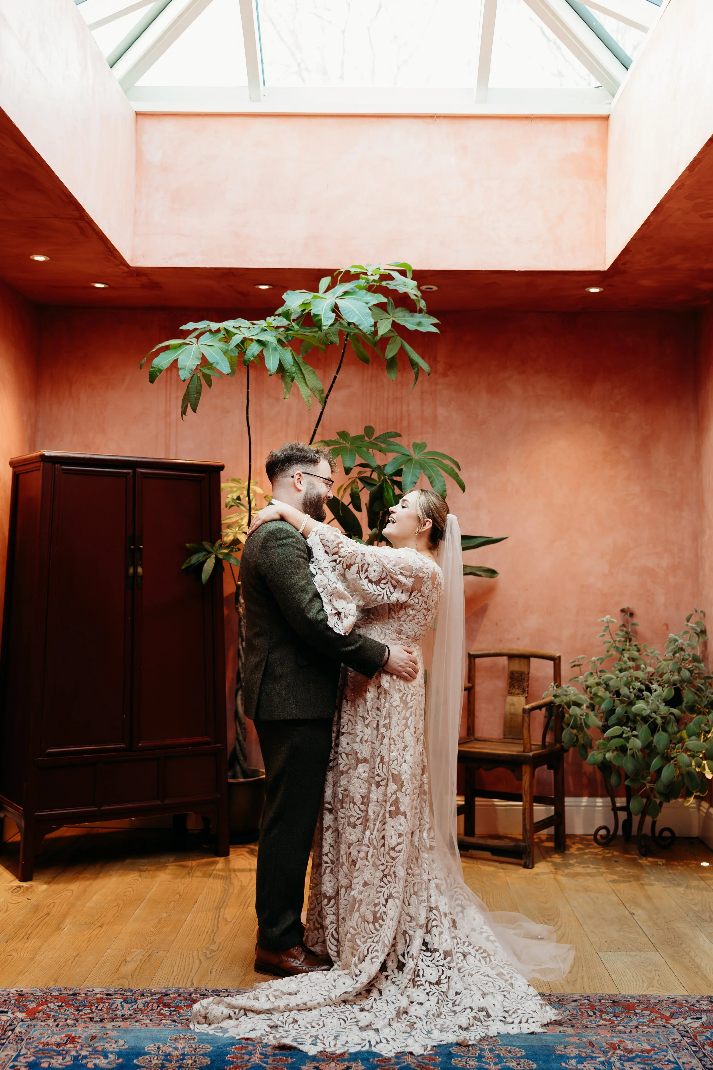 A bride and groom embrace indoors under a skylight with pink walls and tall plants, celebrating their wedding.