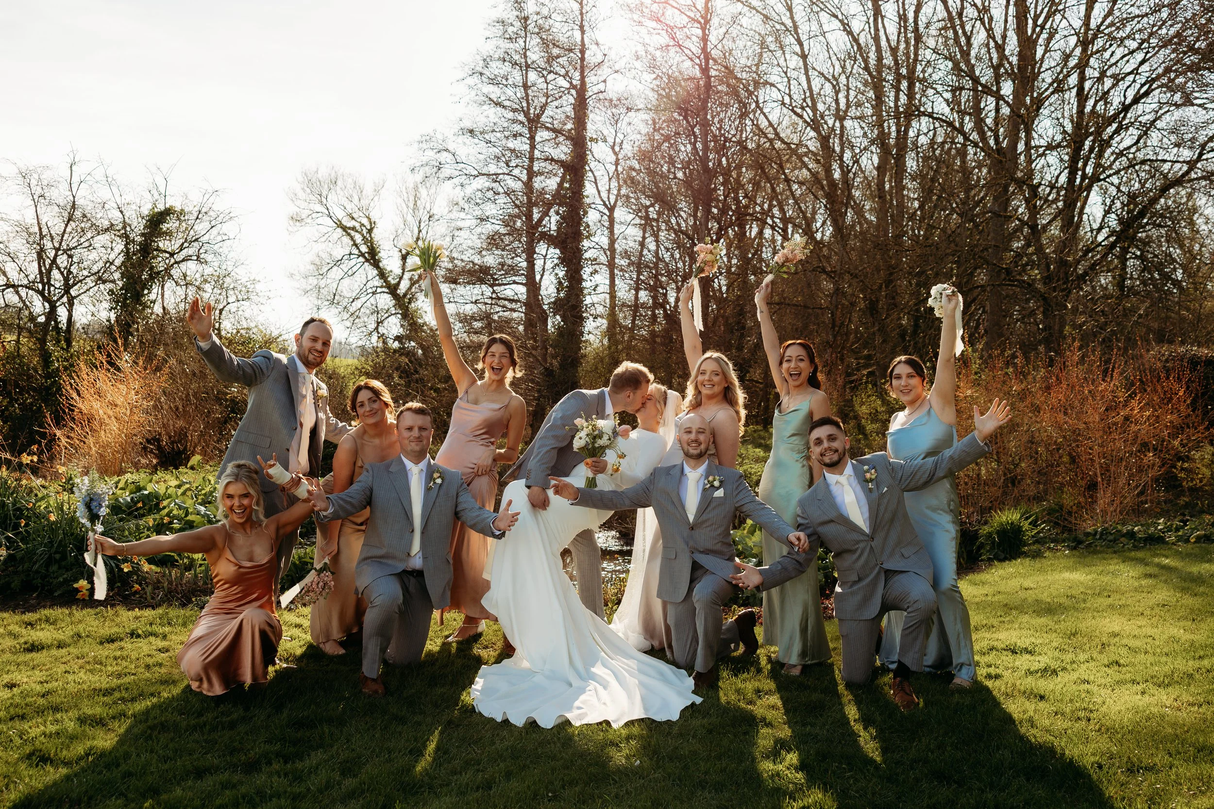 A wedding party outdoors with the bride and groom kissing, surrounded by friends, all celebrating and smiling on a grassy area with trees in the background.