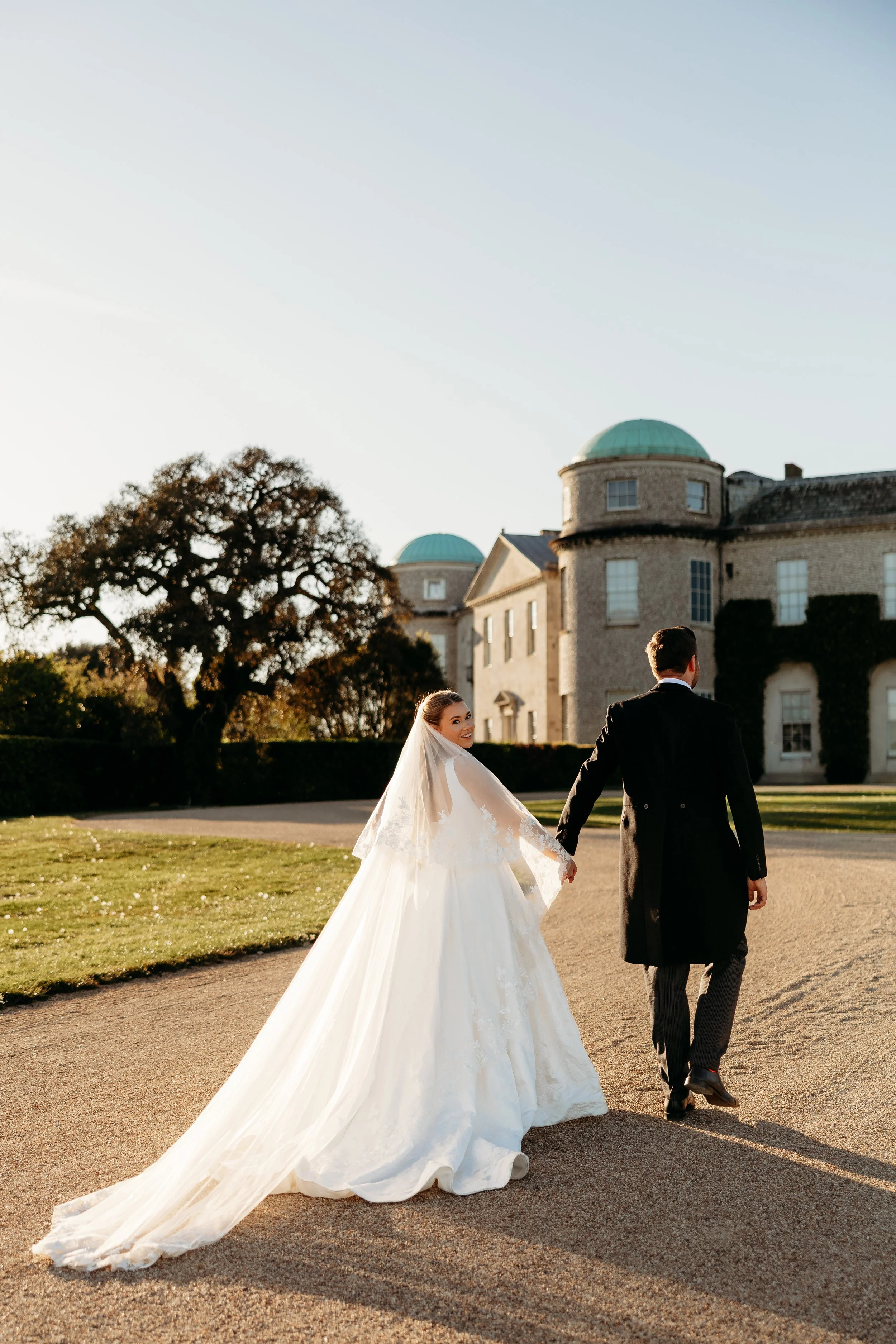 A bride in a white wedding gown with a long train and veil, holding hands with a groom in a black jacket, walking outdoors near a historic building with green domes, during sunset at Goodwood Estate, Sussex.