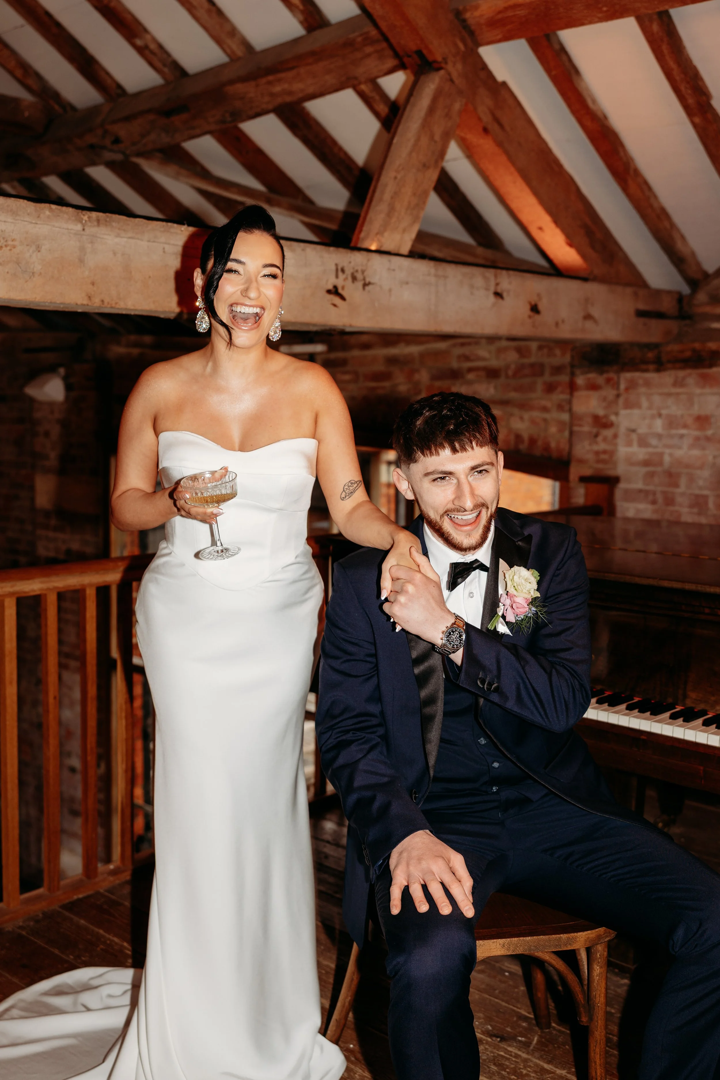 Bride in a white wedding gown and groom in a navy tuxedo with a boutonniere, laughing and smiling at a wedding celebration in a rustic venue with wooden beams and brick walls.