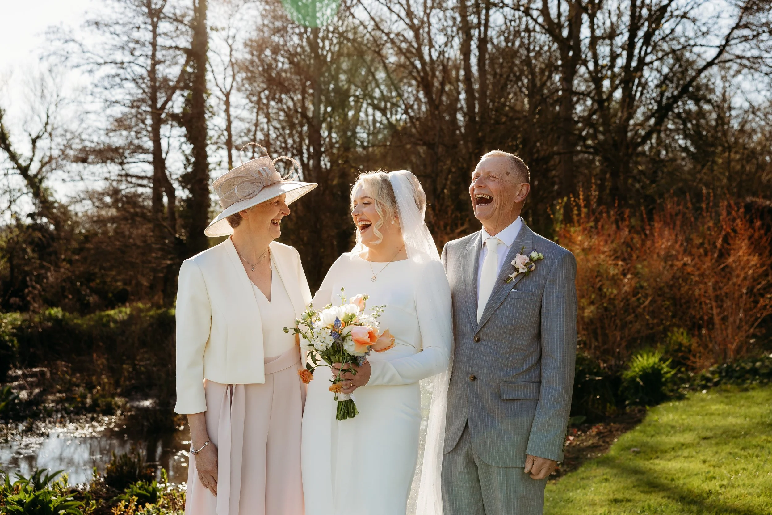 A bride in a white wedding dress holding a bouquet, smiling with her parents on a sunny day outdoors surrounded by trees and greenery.