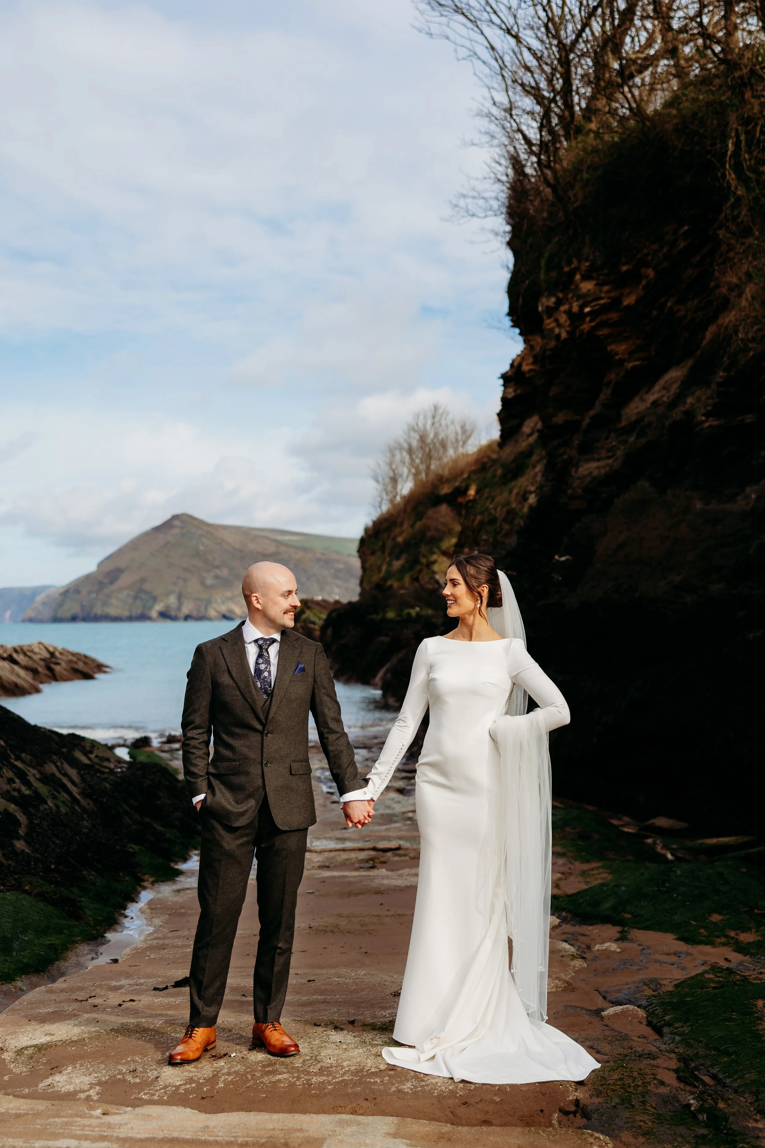 A bride and groom holding hands on a beach with cliffs and ocean in the background.
