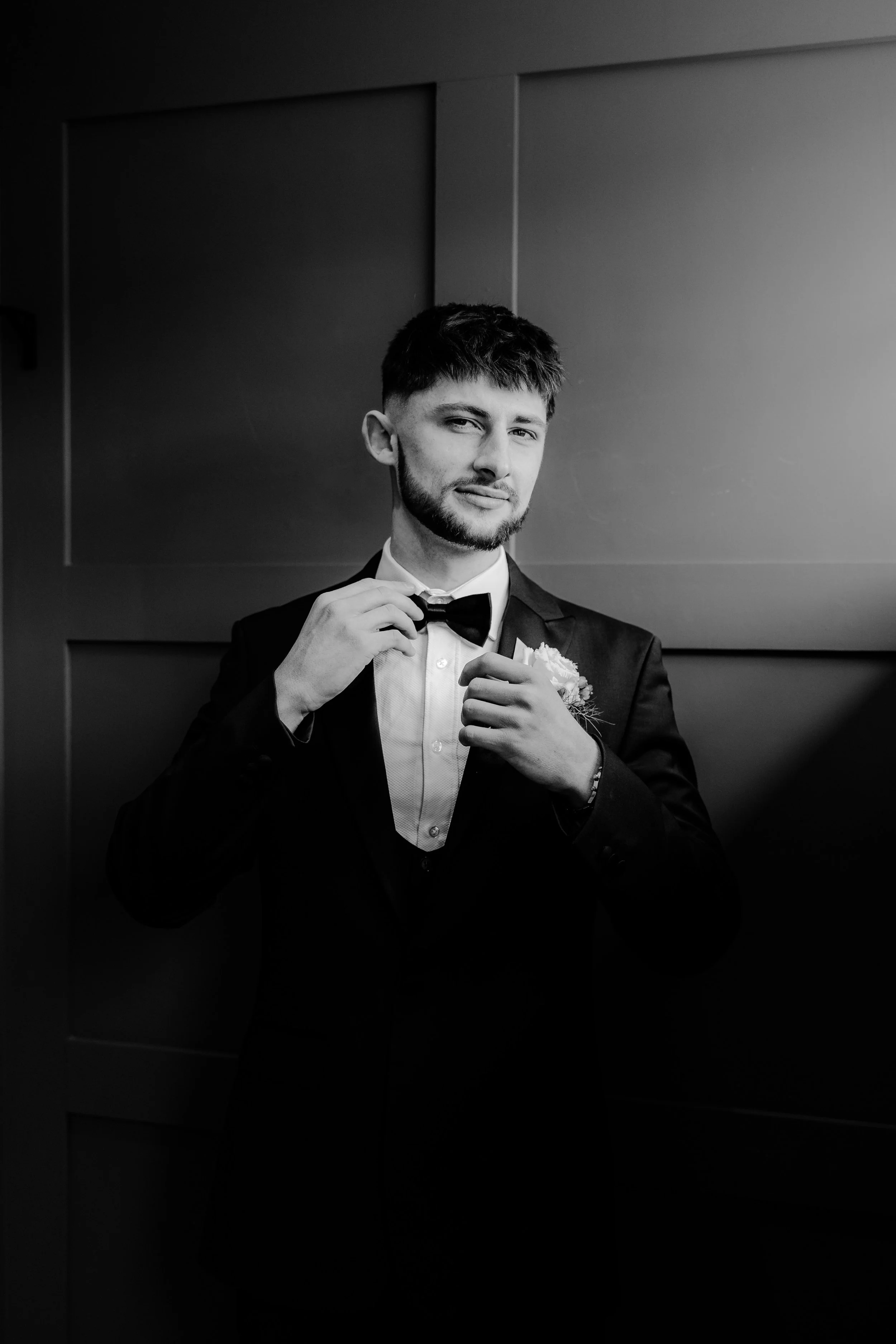 Black and white photo of a man in a tuxedo adjusting his bow tie, standing in front of a dark wall.