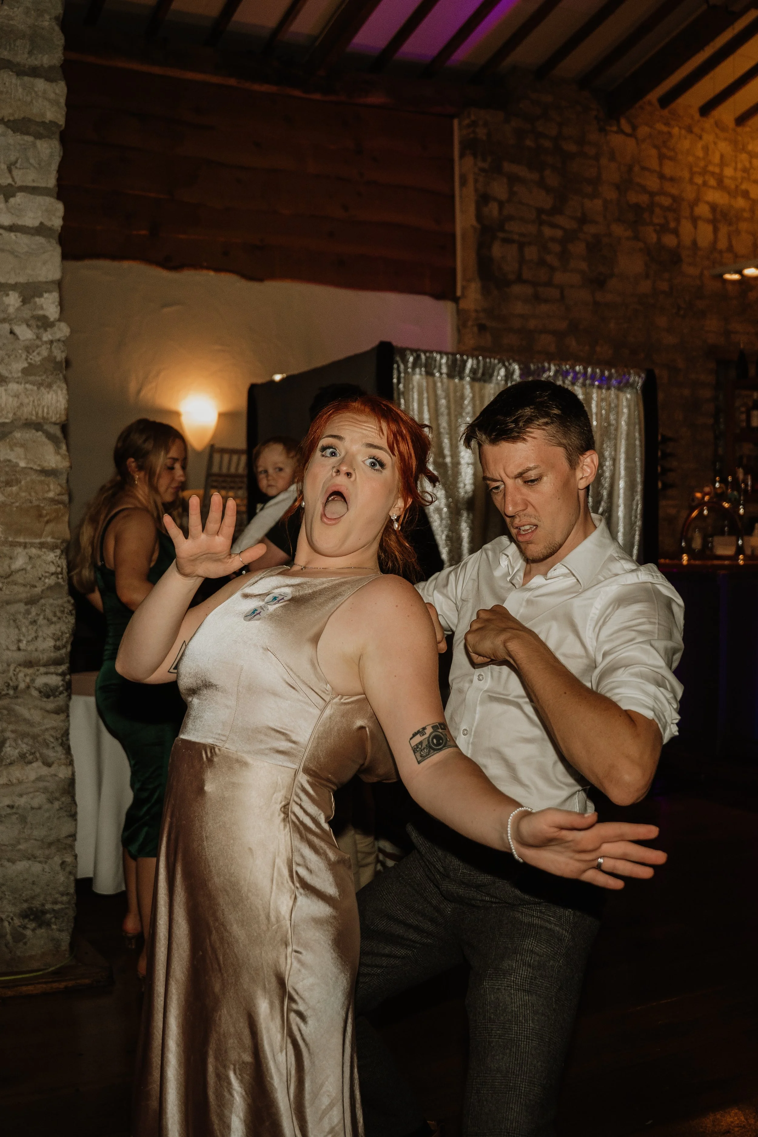 A woman in a gold satin dress with tattoos on her arm and red hair is dancing at a party, with a man in a white shirt dancing close beside her. In the background, other guests are socializing against a rustic brick and wood interior.