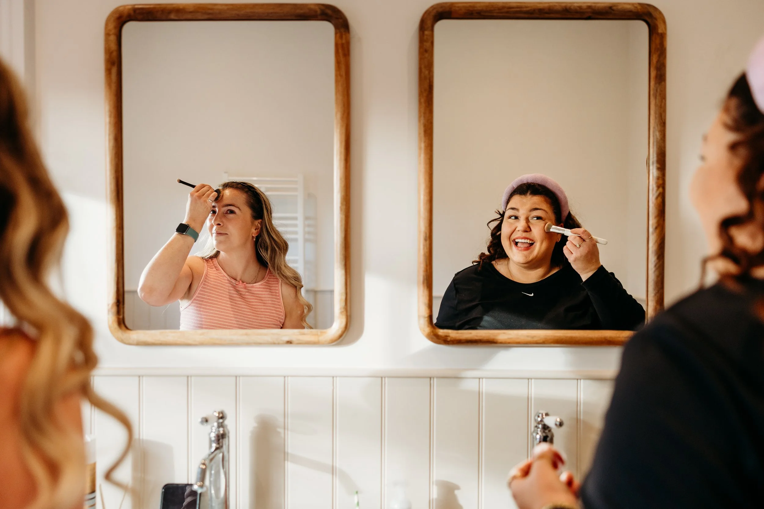 Two women are applying makeup while looking at their reflections in separate bathroom mirrors, one in a pink sleeveless top and the other in a black top with a pink headband.
