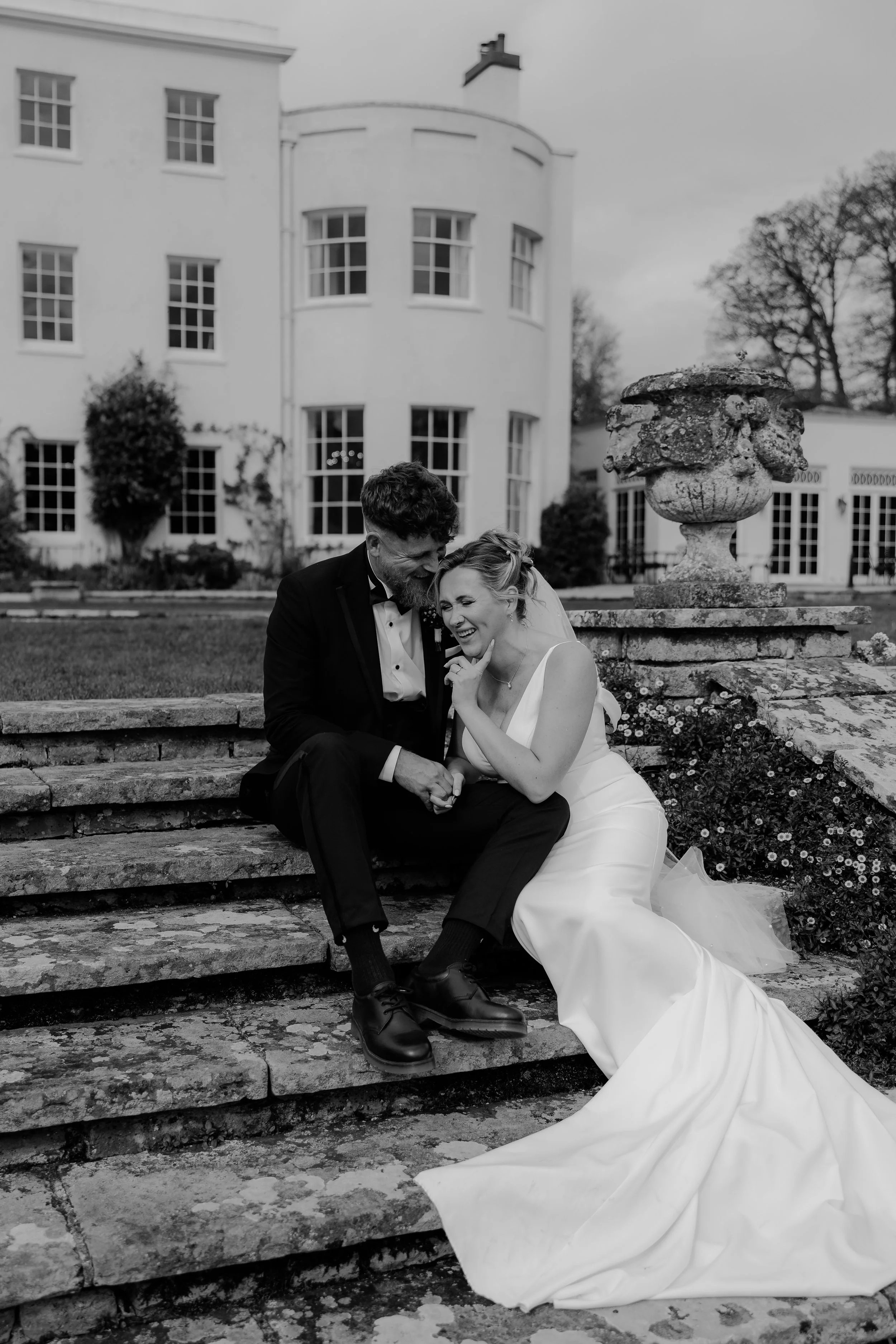 A black-and-white photo of a bride and groom sitting on outdoor stone steps, smiling and holding hands, with elegant architecture and trees in the background.