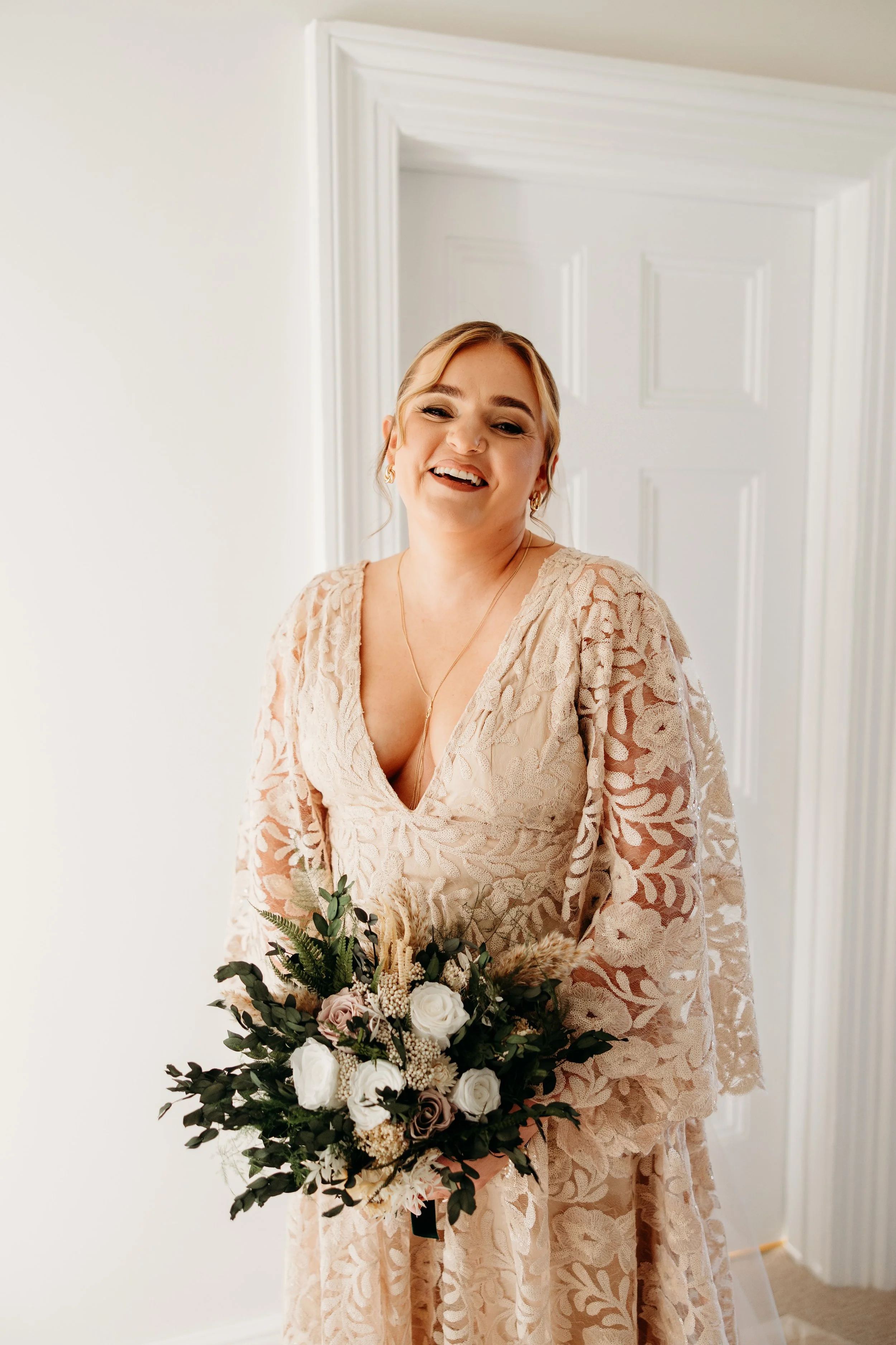 A smiling woman in a lace dress holding a bouquet of flowers, standing in a bright room looking beautiful before her wedding at Matara, Cotswolds.