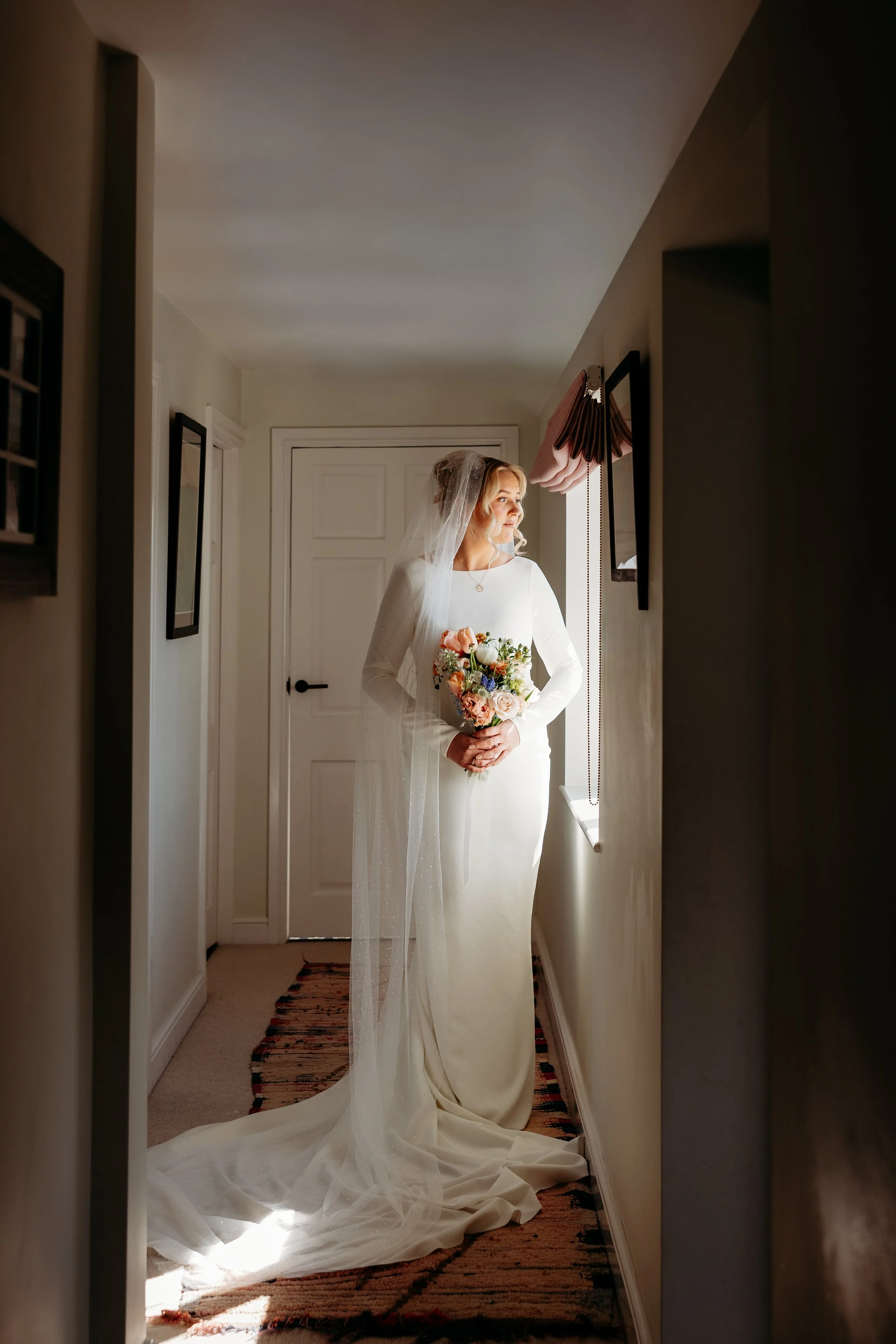 A bride in a white wedding gown and veil holding a bouquet, standing by a window and looking outside in a hallway with beige walls and a patterned rug.