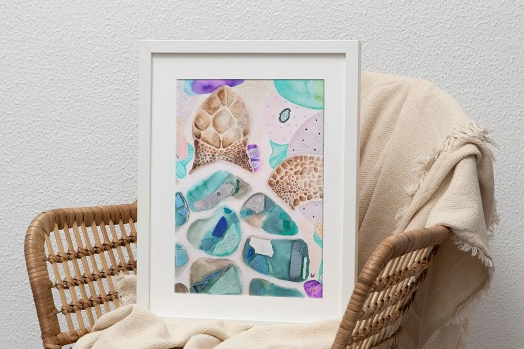Watercolour turtle artwork featuring a variety of abstract stone-like shapes in pastel and vibrant colors, framed and leaning against a textured white wall with a beige cloth draped over the chair.