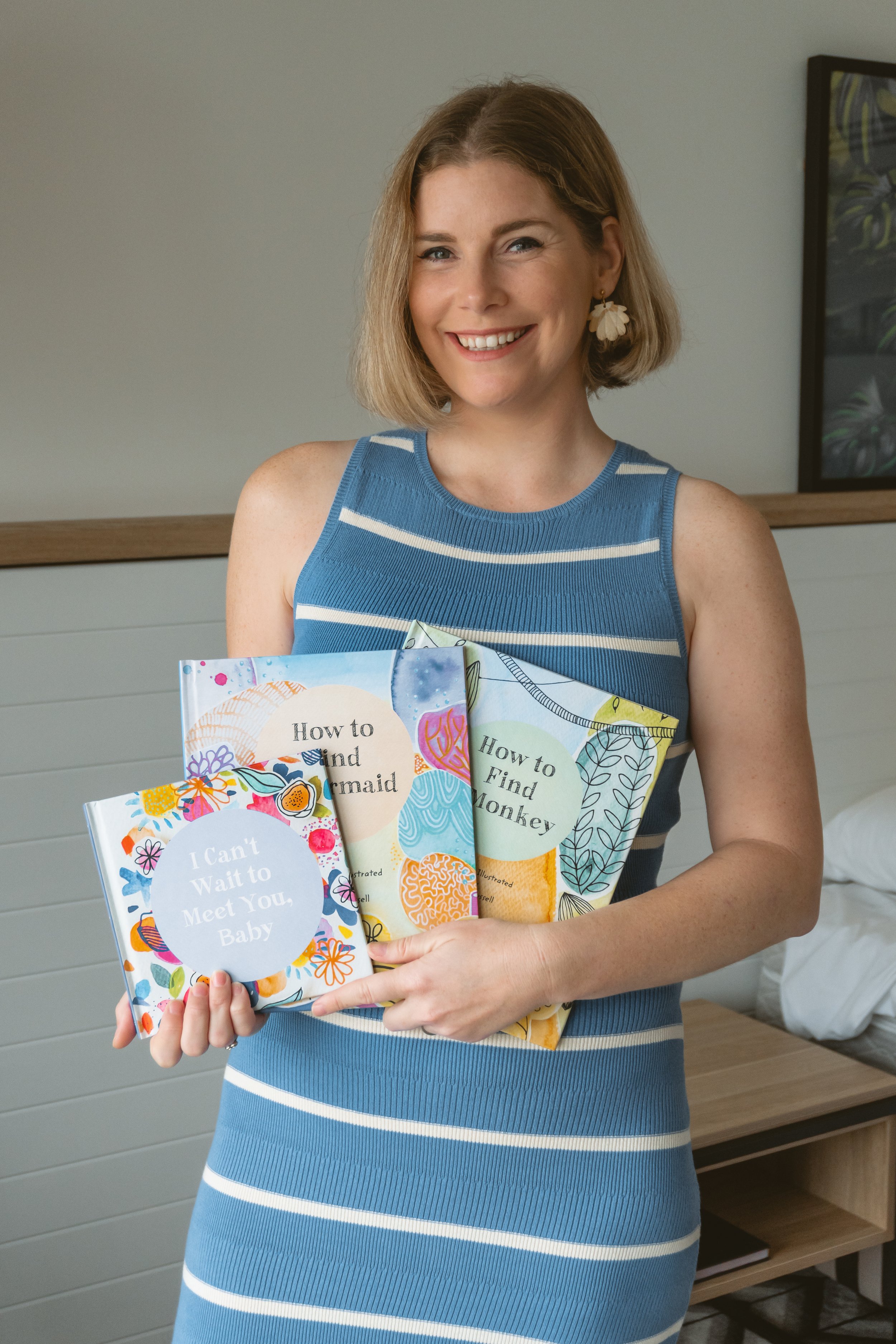 A woman in a blue and white striped sleeveless dress is smiling and holding three colorful children's books titled 'I Can't Wait to Meet You, Baby', 'How to Find Mermaid', and 'How to Find Monkey' in a room with beige walls and a bed.
