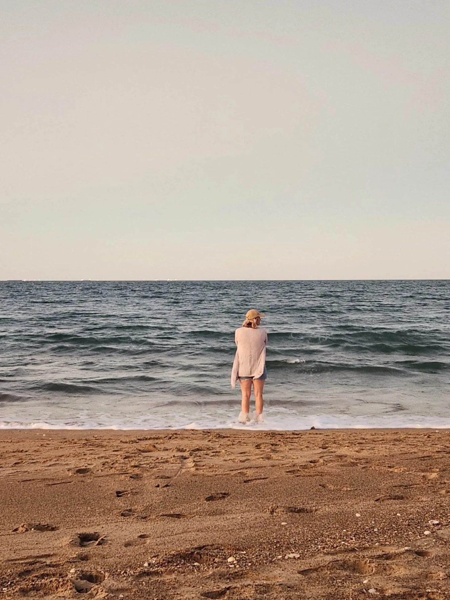 A woman standing at the edge of the ocean on the beach, facing the water, wearing a hat, a light-colored long-sleeve shirt, and shorts.