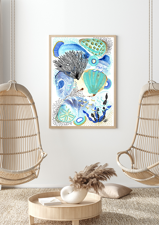 A framed abstract ocean-themed artwork hangs on a white wall between two hanging rattan chairs. A round wooden coffee table in front holds a white vase with pampas grass and a book. A beige cushion is on the floor beside a decorative pillow.