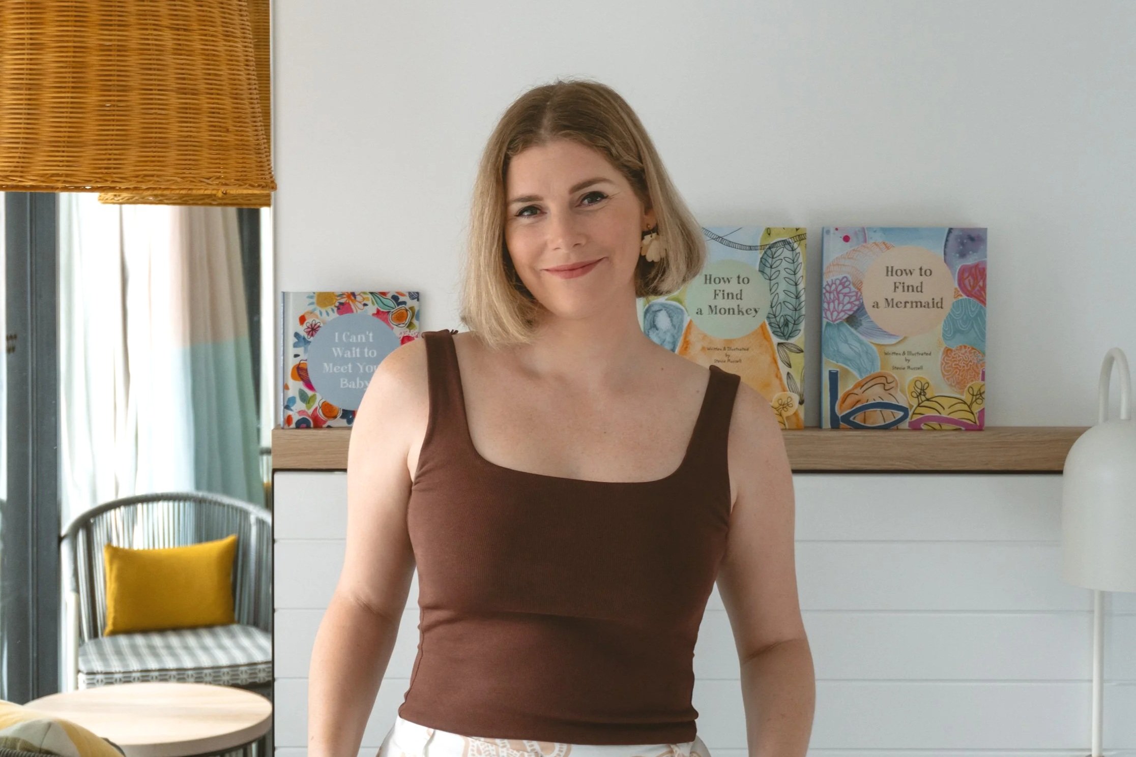 A woman with shoulder-length blonde hair and a brown tank top smiling at the camera in a bright room with books and a chair in the background.