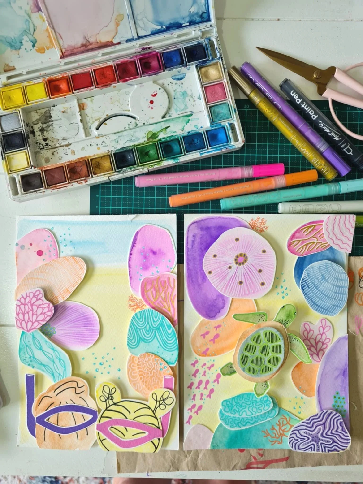 Watercolour art supplies and completed watercolor artworks featuring colorful abstract and floral designs, with watercolour paints, brushes, markers, and scissors on a workspace.