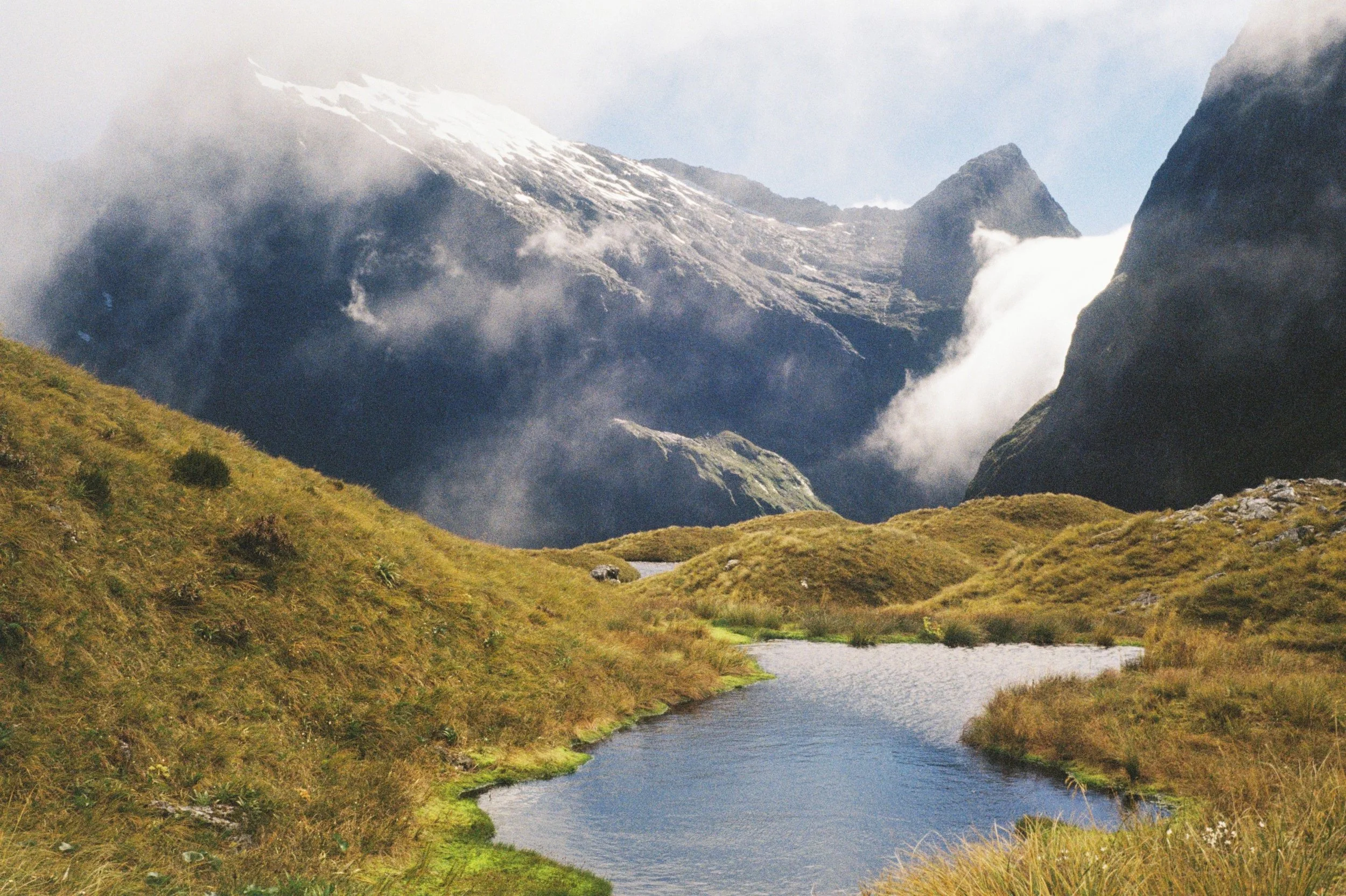 A scenic landscape featuring a river flowing through lush green grass with mountain peaks in the background, partially covered in snow and surrounded by clouds.