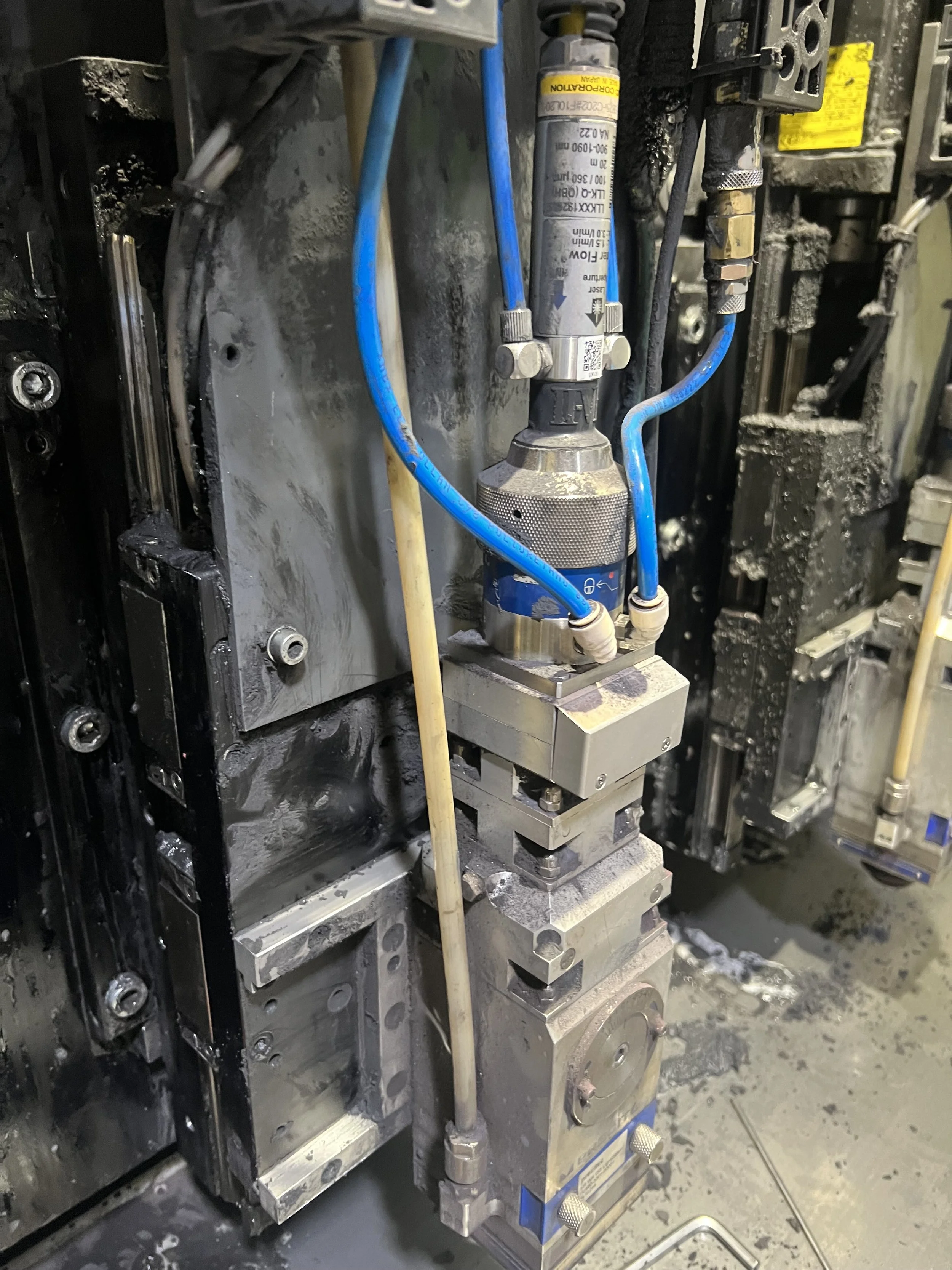 Close-up of industrial machinery with hydraulic components, blue and yellow hoses, and metal parts, showing signs of use and oil or coolant residue.