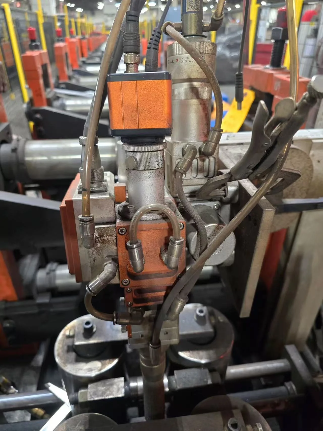 Close-up of mechanical equipment and tubing, with orange-colored components, in an industrial setting, possibly part of a manufacturing or machining process.