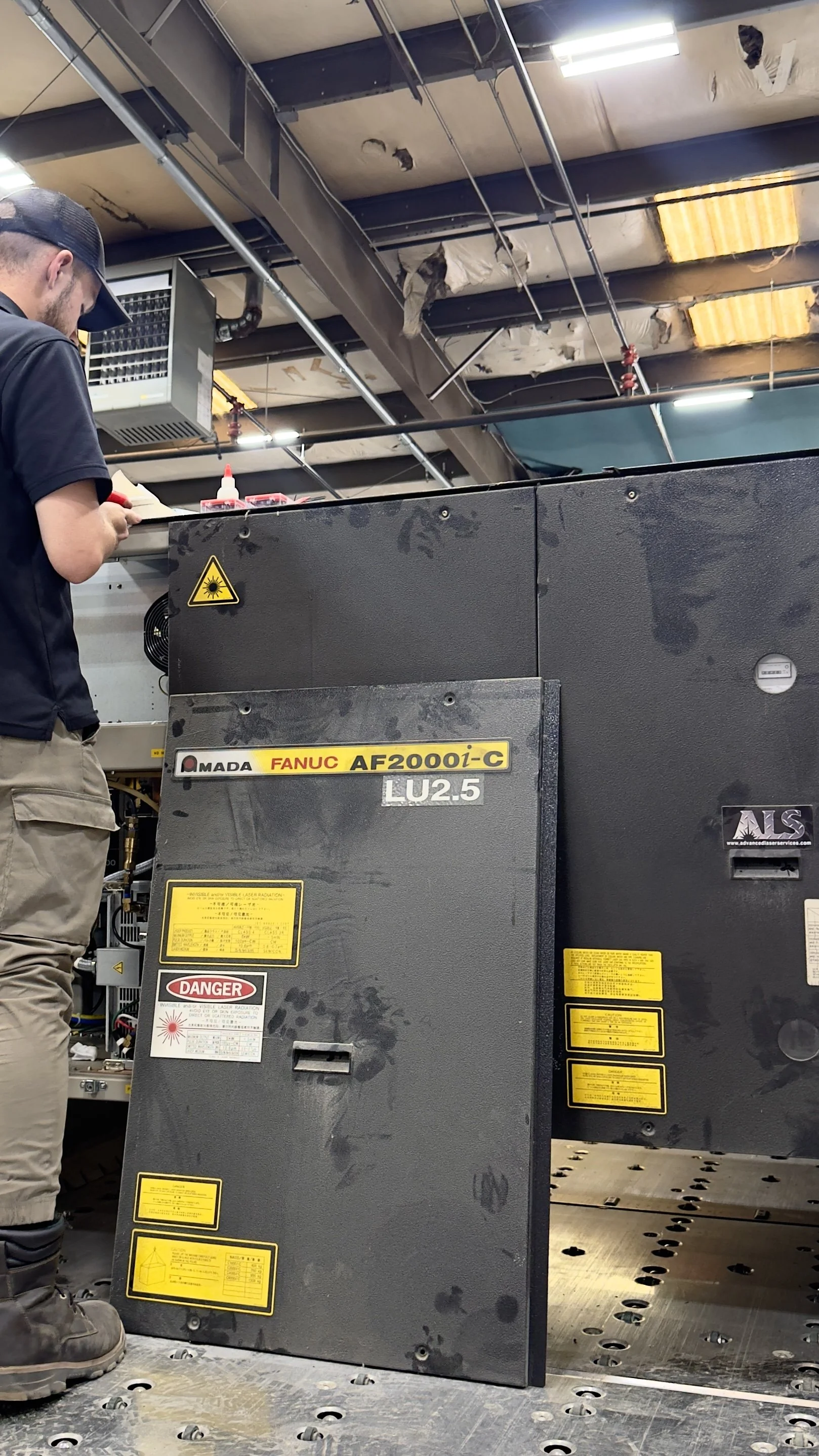 A technician standing next to a large industrial laser machine in a factory setting. The laser machine has yellow and black warning labels, including a caution sign for laser radiation. The ceiling is damaged with torn insulation and exposed beams.