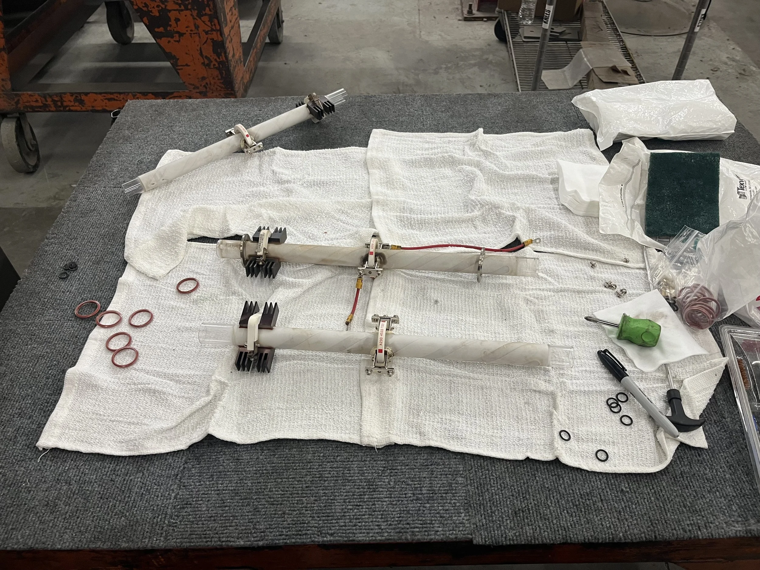 Disassembled antenna components laid out on a white cloth, including long plastic pipes with metal clamps, O-rings, a green screwdriver, and various small parts and tools for assembly or repair.
