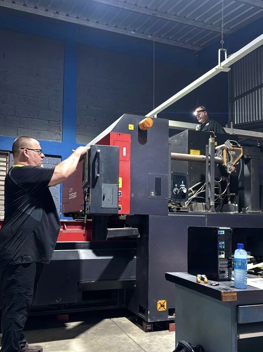 Two men working on a large industrial machine inside a warehouse. One man is standing and wearing glasses, operating or inspecting the machine, while the other man is on the platform above, also wearing glasses and safety equipment, smiling.