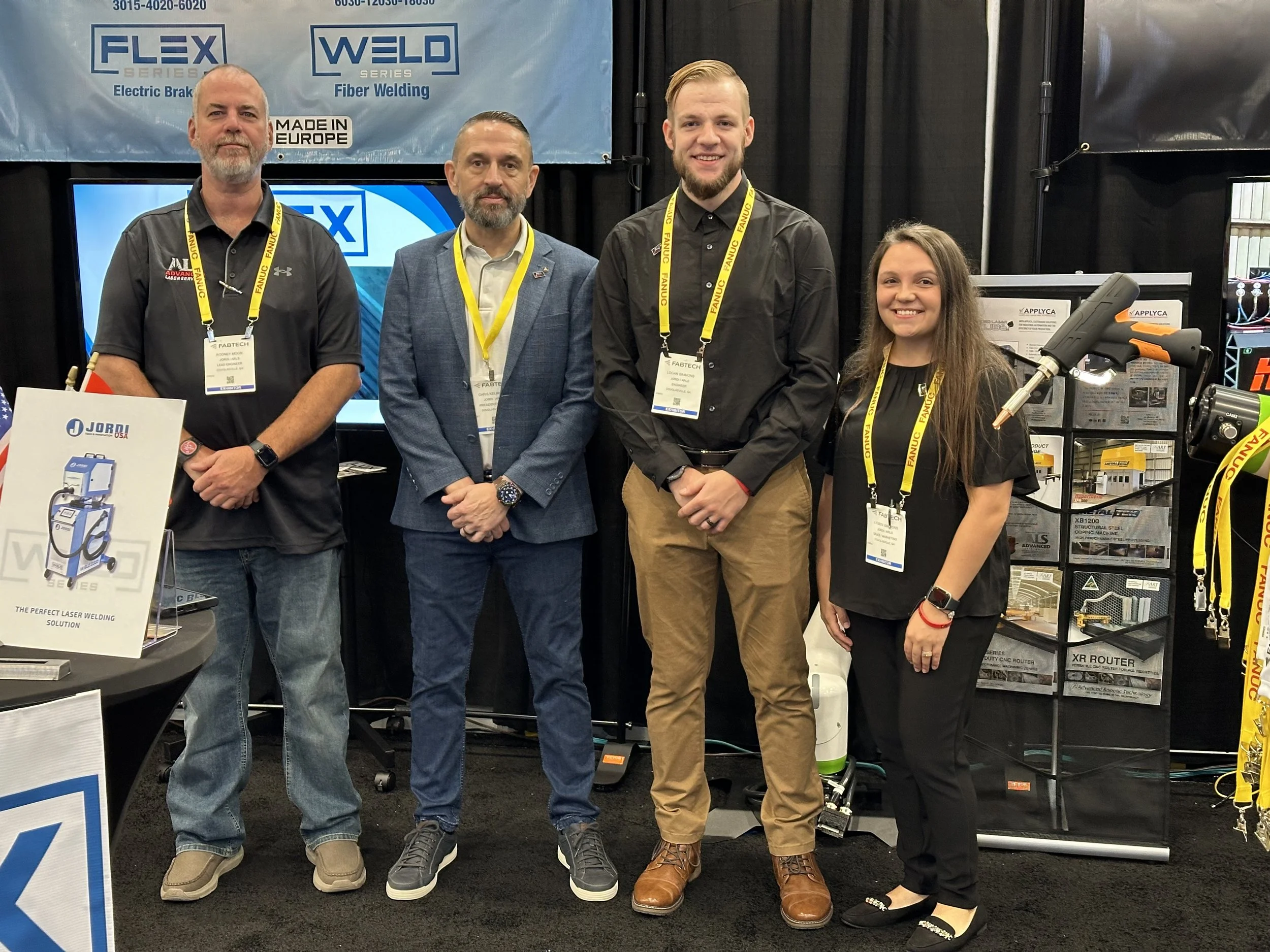 Four people posing at a trade show booth related to fiber welding and electric brakes, with banners displaying 'FLEX Series' and 'WELD Series'.