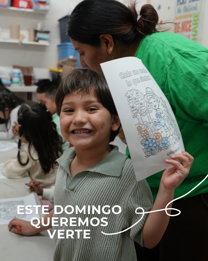 Un niño sonriendo sosteniendo un dibujo de una niña en un jardín con flores. En el fondo, están otros niños en lo que parece ser una actividad escolar en un aula.