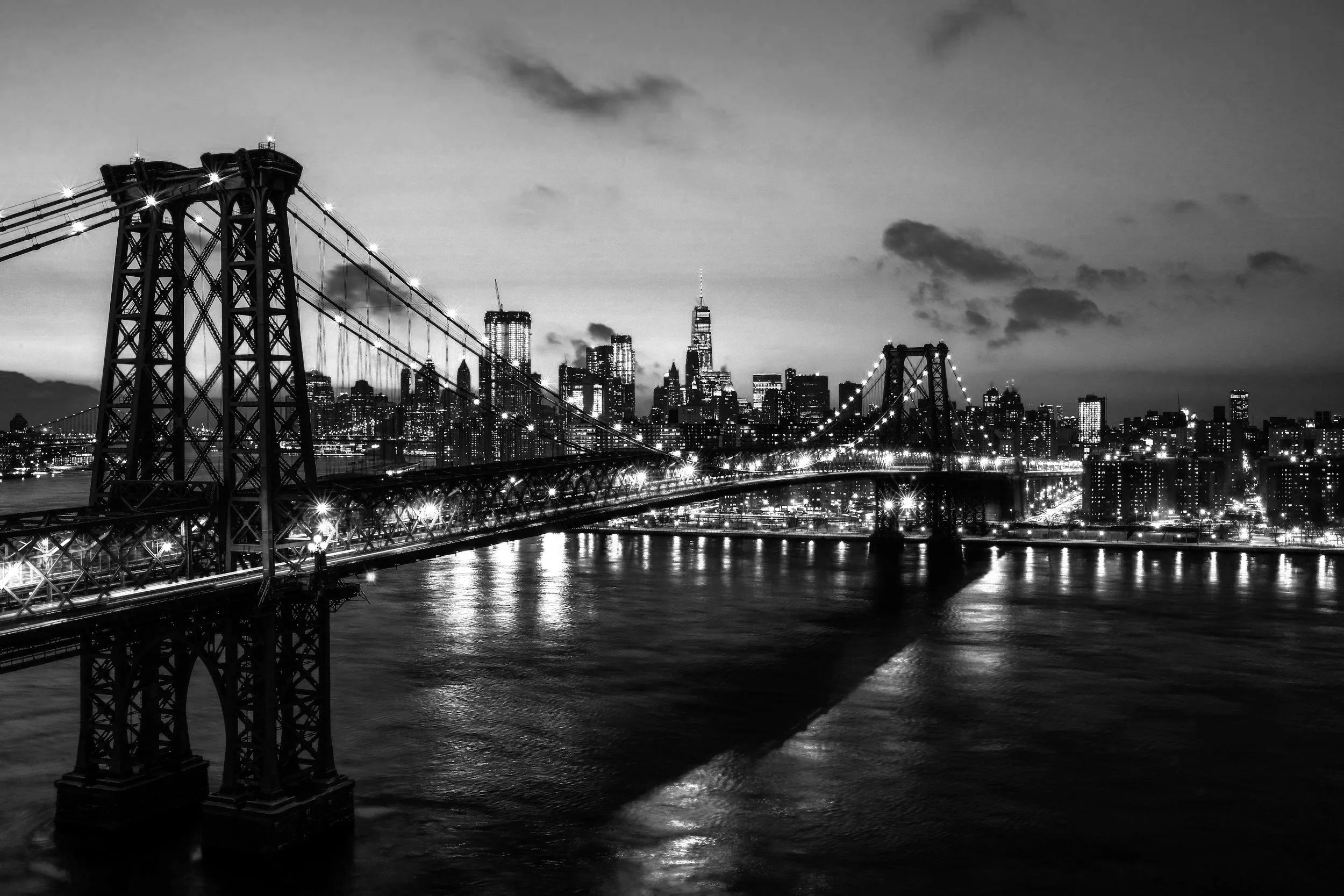 Puente de Brooklyn iluminado en la noche con la ciudad de Nueva York de fondo en un cielo nublado y oscuro.