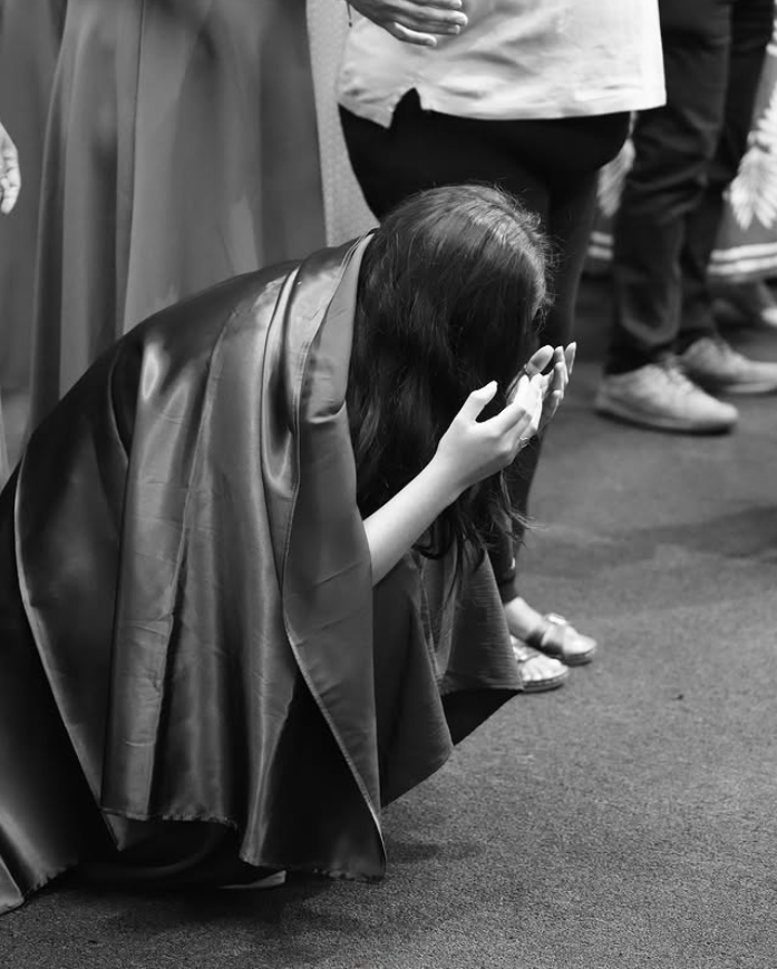 Una mujer arrodillada con ropa de ceremonia, cubriéndose el rostro con las manos, en un espacio cerrado con varias personas de pie alrededor.