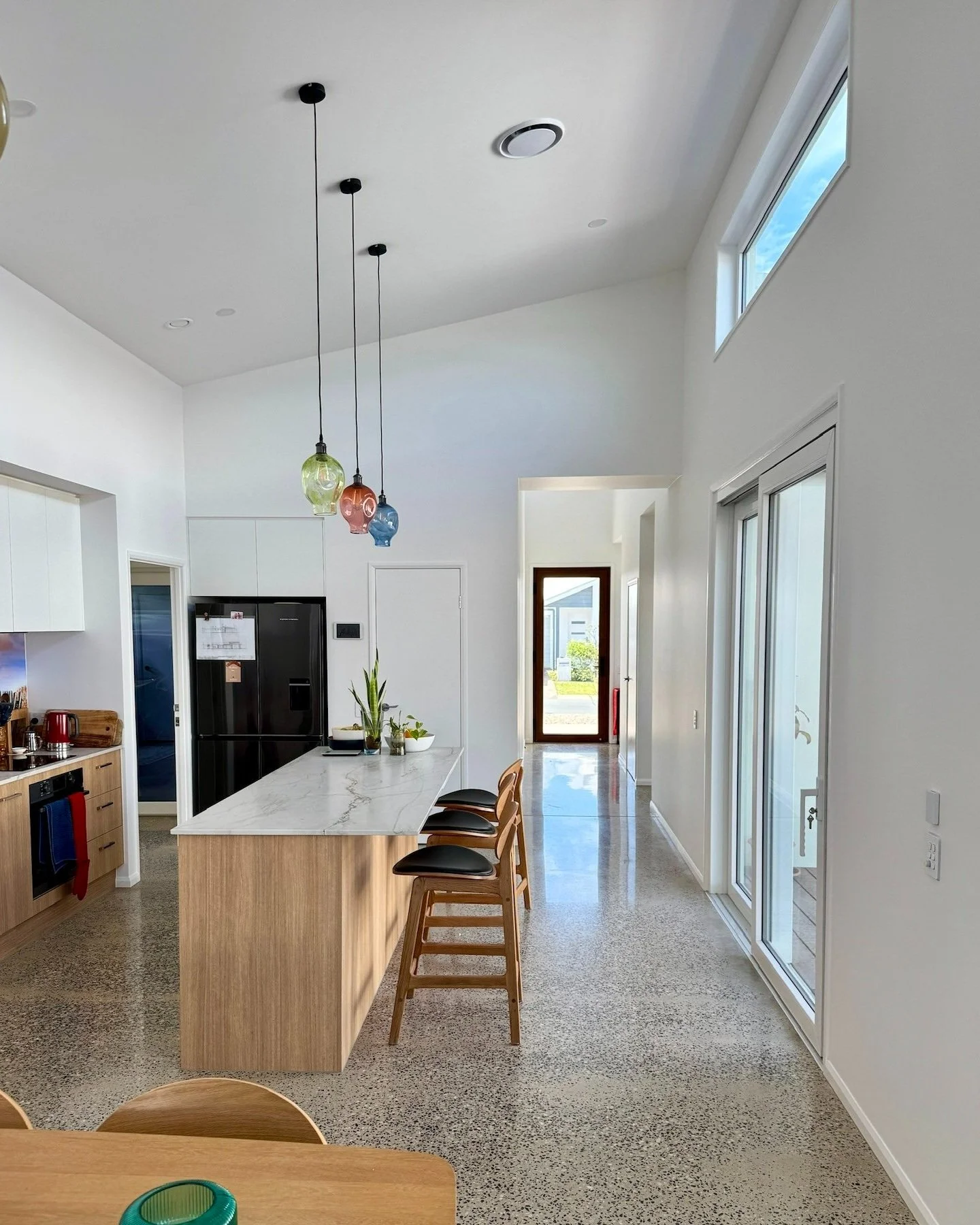Modern kitchen with white walls, a marble island, colorful pendant lights, and sliding glass doors leading outside.