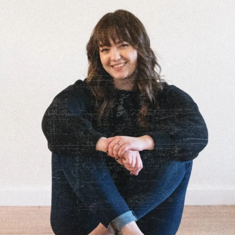 A smiling woman with wavy brown hair and bangs sitting on the floor against a plain white wall, wearing a black sweater and blue jeans.