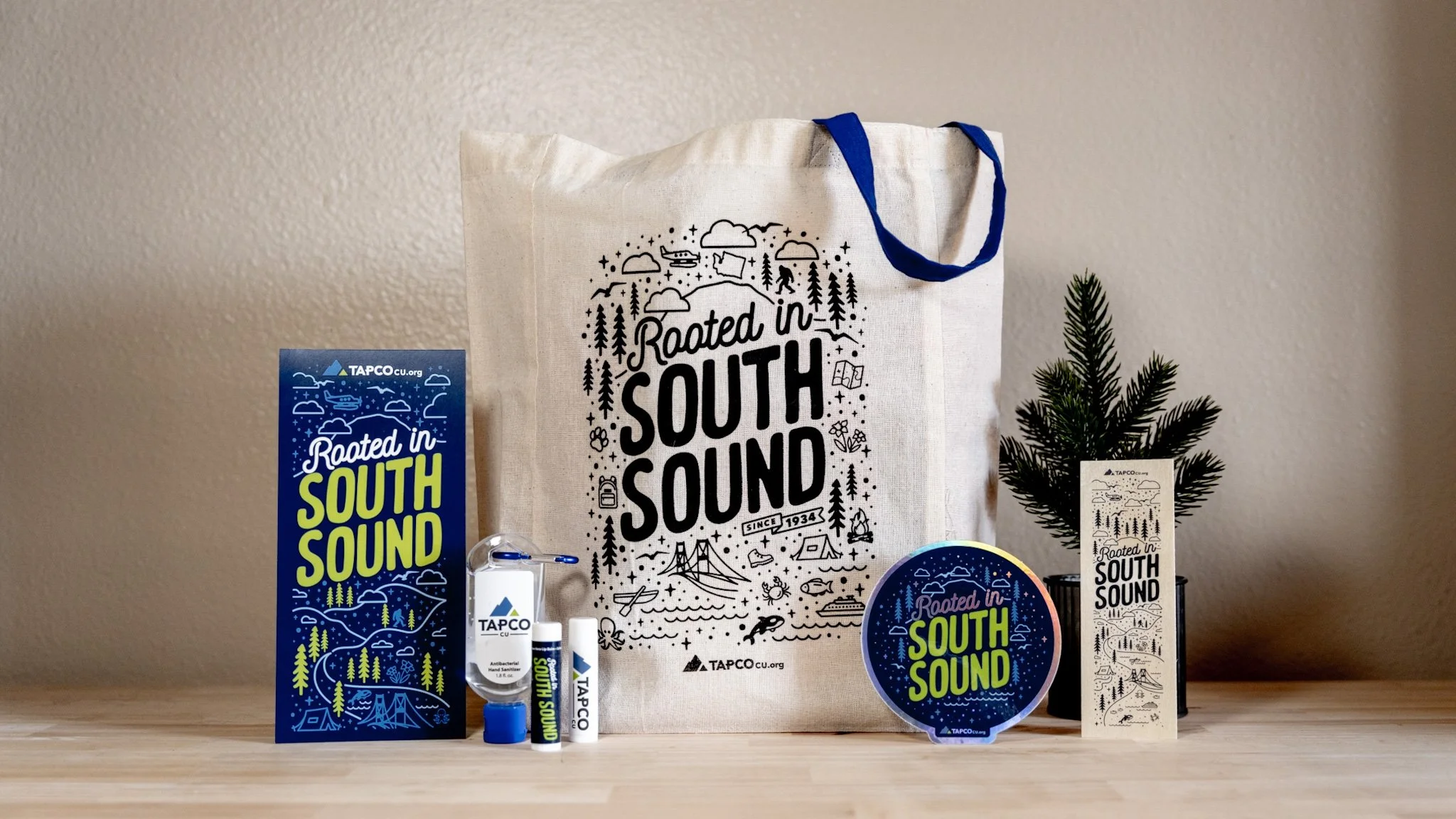 Various promotional items and merchandise for 'Rooted in South Sound,' including tote bags, stickers, and hand sanitizer, displayed on a light-colored wooden surface against a beige wall. A small artificial pine tree is also present.