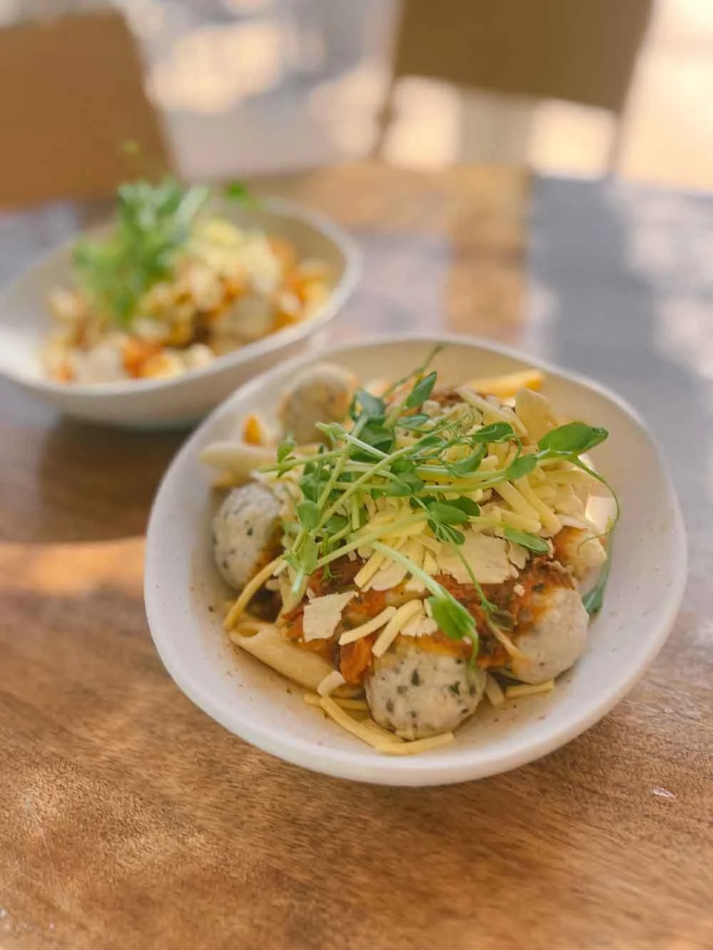 Bowls of noodles topped with shredded cheese, microgreens, and meatballs on a wooden table.