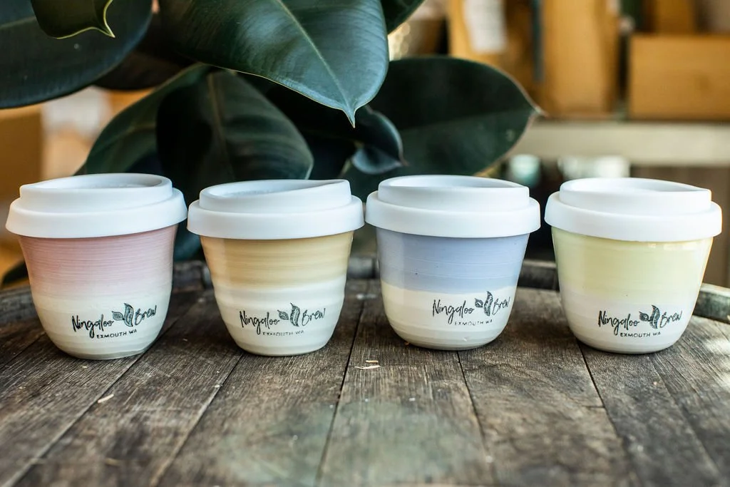 Four ceramic cups with white lids and pastel-colored gradients, labeled with 'Ningaloo Brew' on a wooden table, with green leafy plant in background.