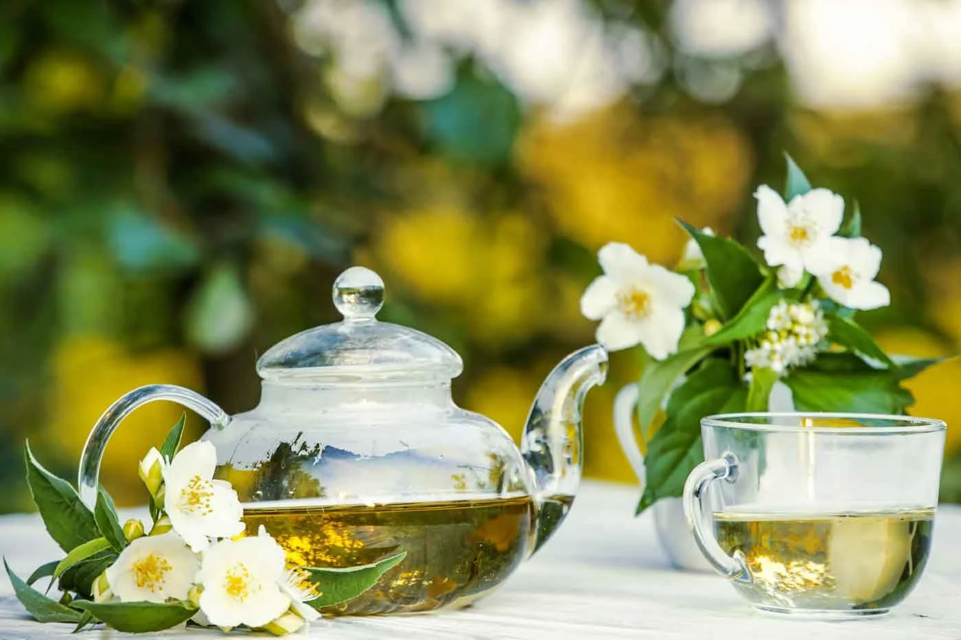 A glass teapot and cup filled with tea on a white table, surrounded by white flowers and green leaves, outdoors with a blurred background.