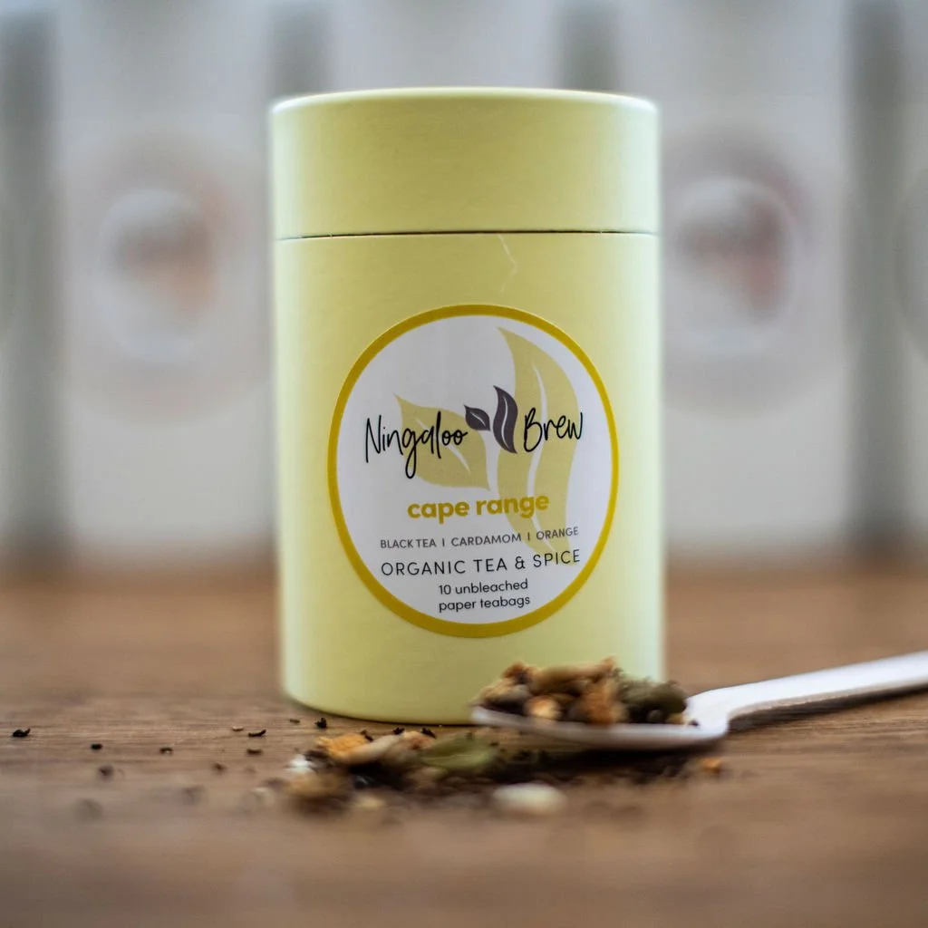 Yellow tin container of Ningaloo Brew Cape Range organic tea with a spoon of loose tea leaves and peppercorns in front, blurred background.