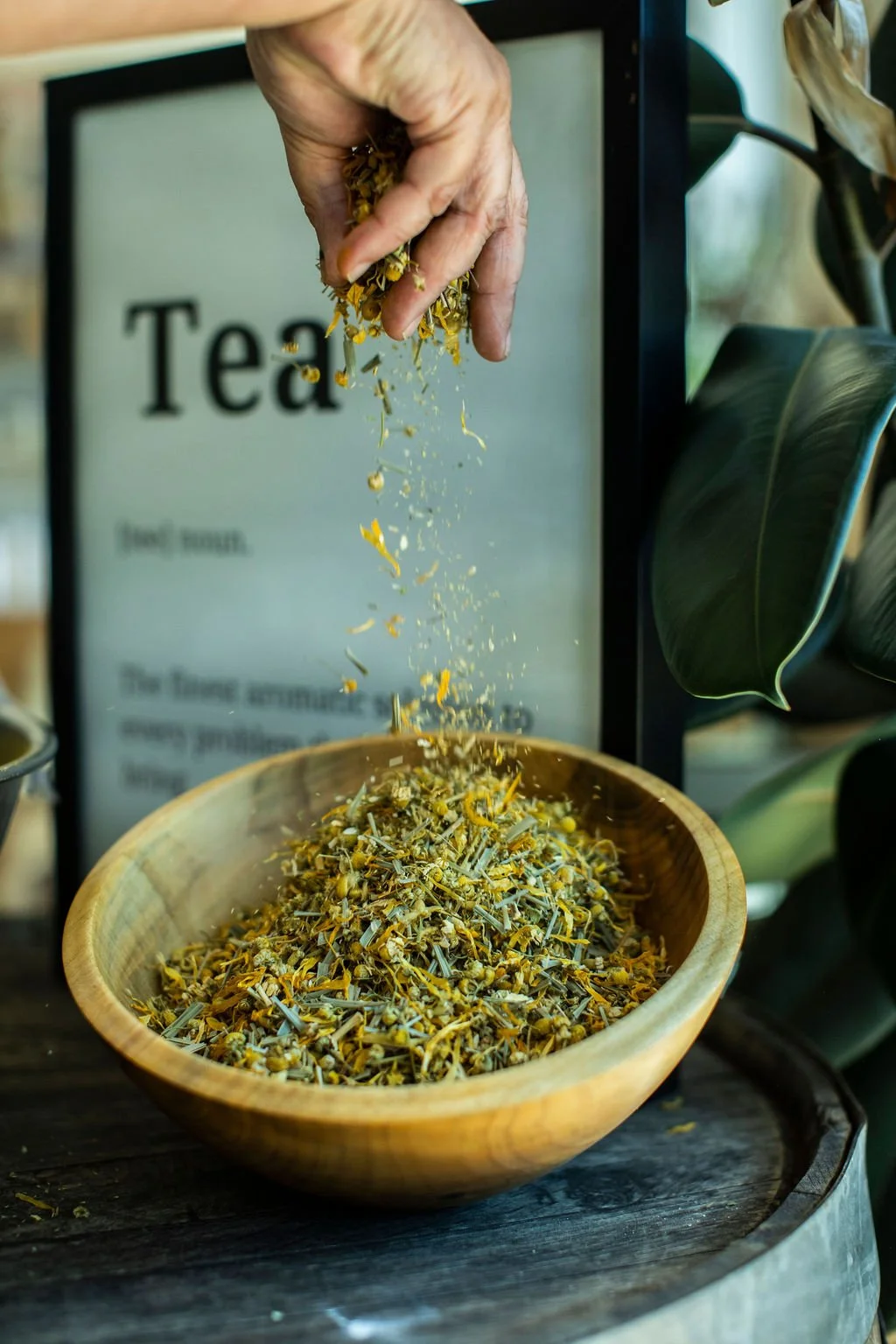 A hand sprinkling dried herbal tea leaves into a wooden bowl, with a sign in the background indicating 'Tea'.